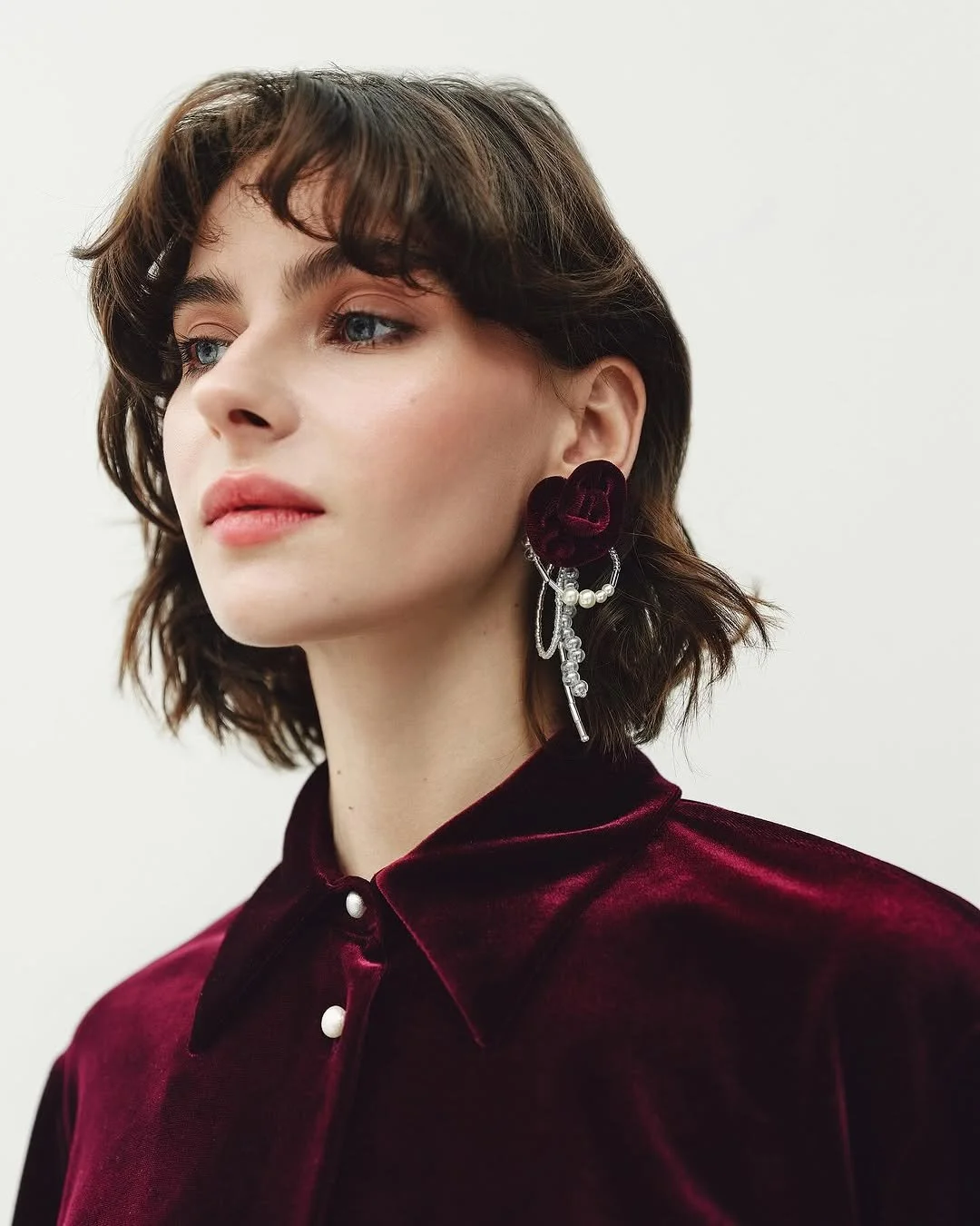 Woman wearing a burgundy velvet shirt with pearl buttons and large statement earrings, featuring pearls and floral designs, against a white backdrop.