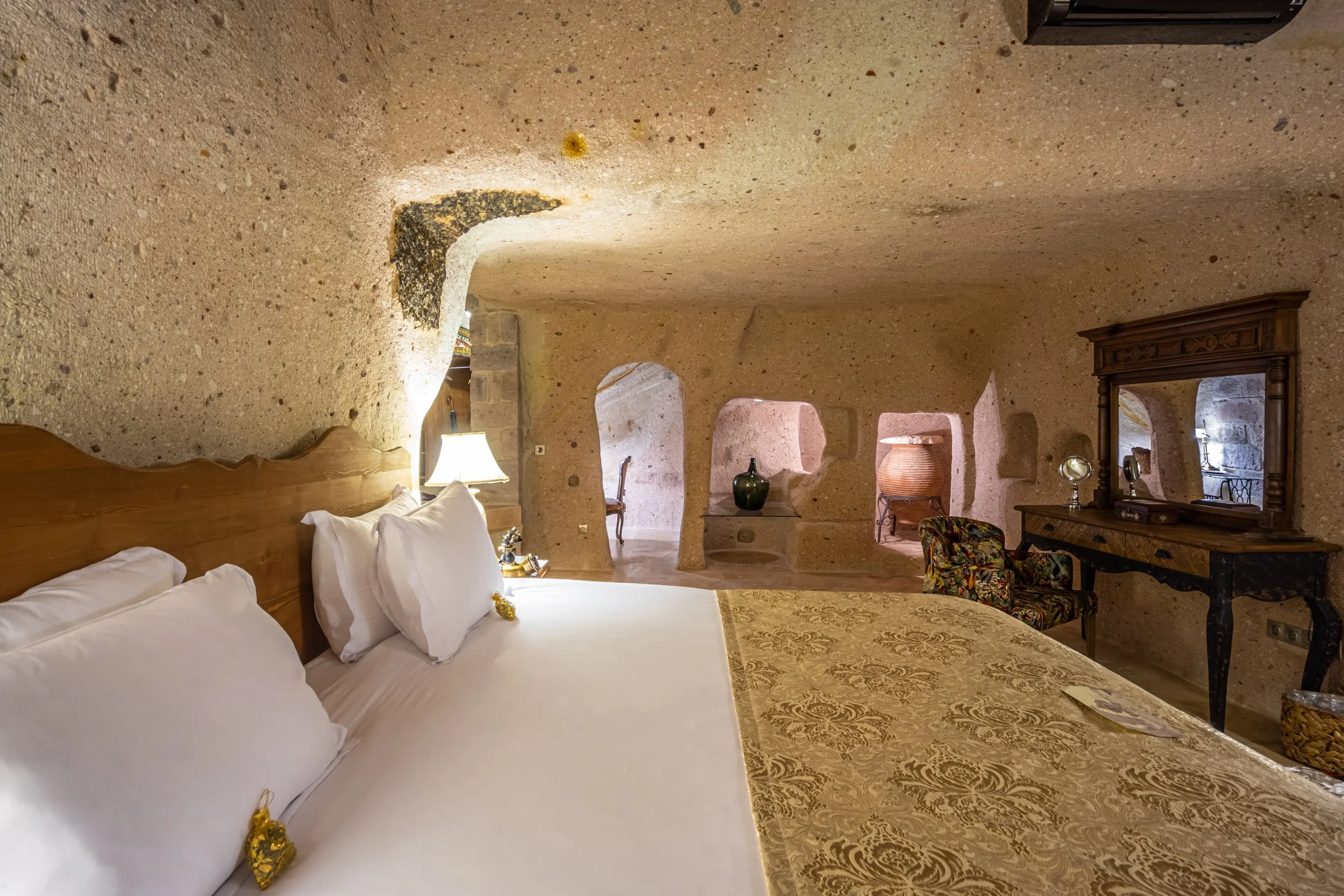 Luxury cave hotel room with a large bed, pillows, ornate bedding, stone walls, wooden dresser, mirror, and decorative vases.