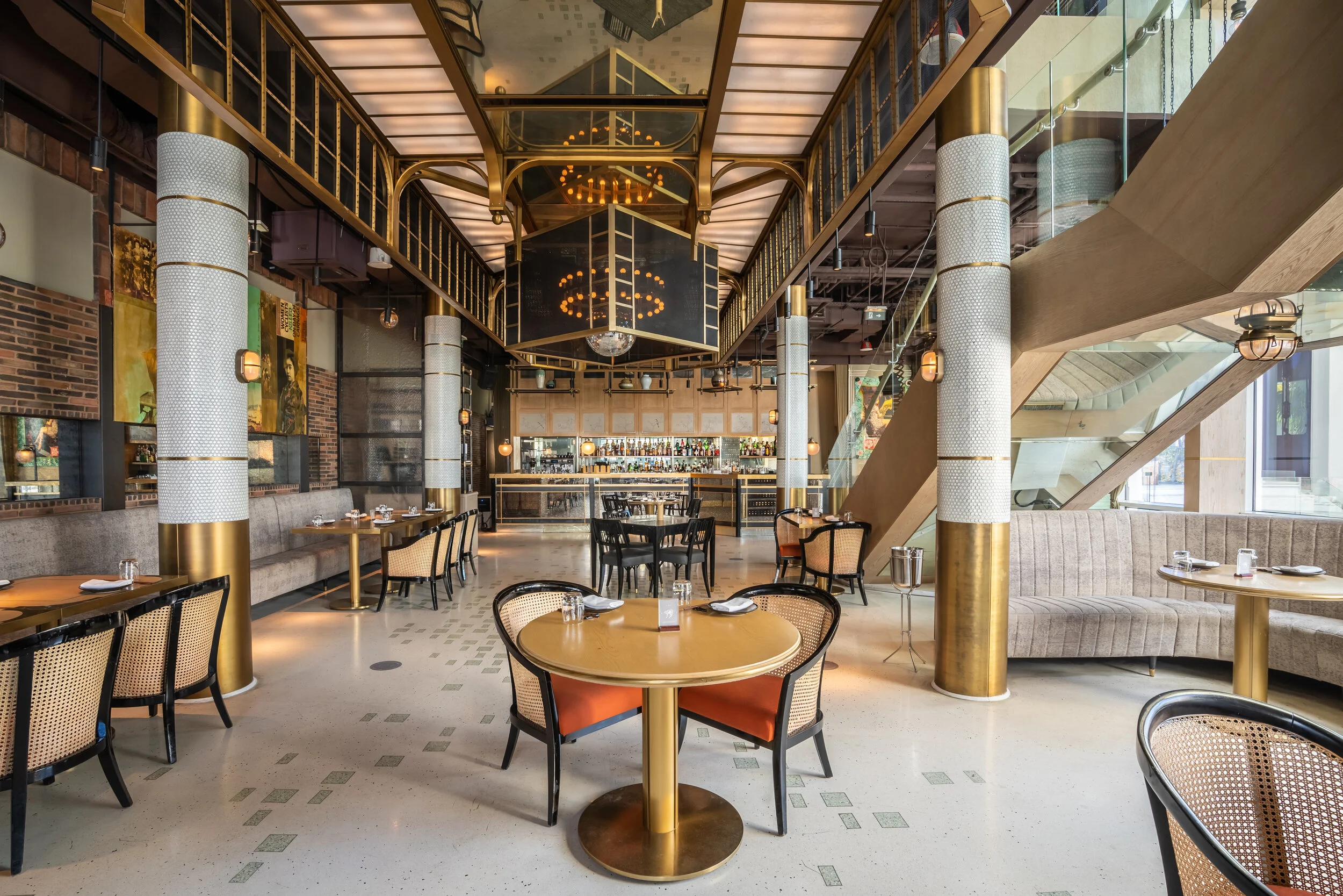Upscale restaurant interior with modern decor, featuring round tables and chairs, elegant lighting, and a stylish bar area in the background. The space includes large pillars and a staircase.