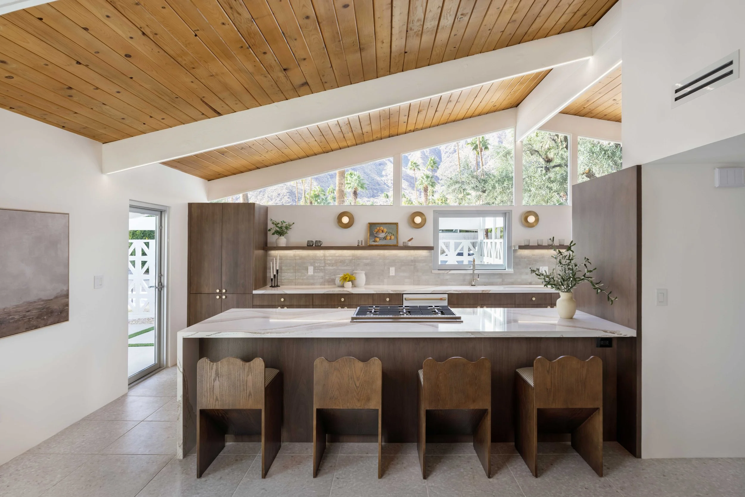 Modern kitchen with a wooden ceiling, marble countertop island, four wooden bar stools, brown cabinetry, and large windows.