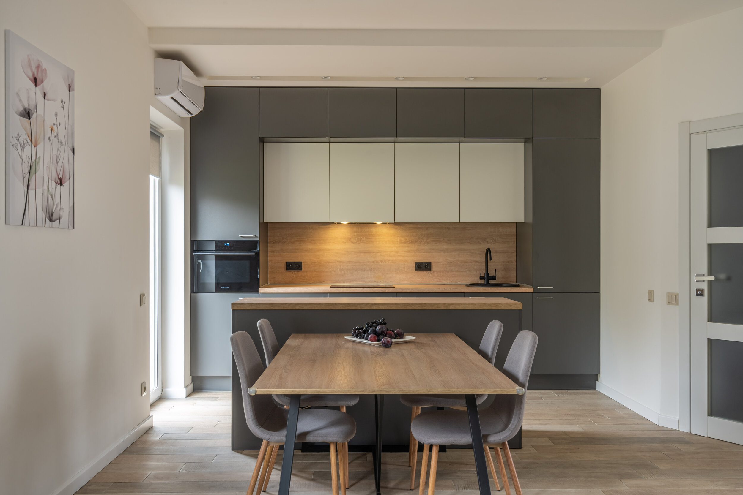 Modern kitchen with gray cabinetry, a wooden table and chairs, built-in appliances, and a floral wall painting.