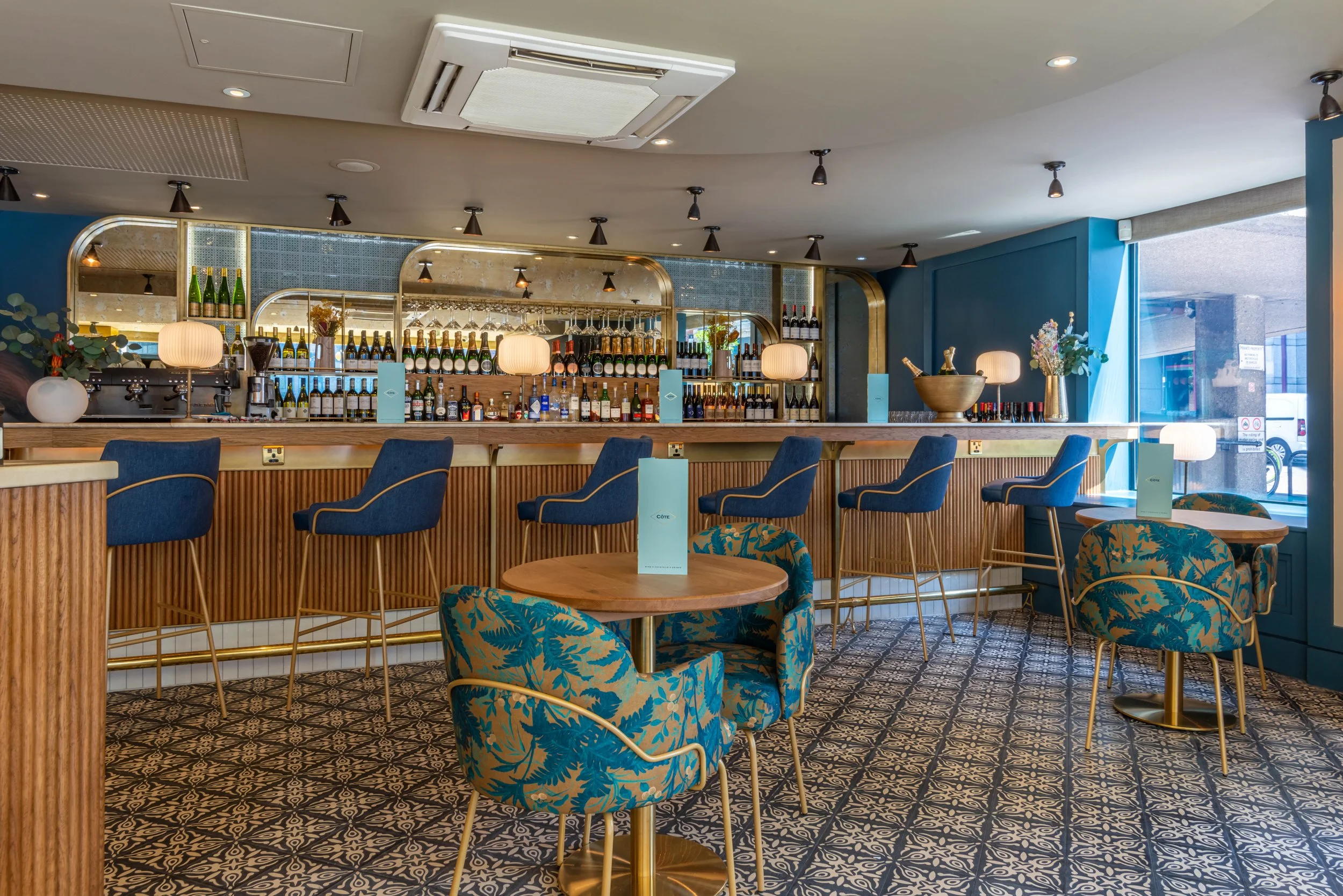 Modern bar interior with patterned tile floor, wooden bar, blue and gold bar stools, round tables with teal and gold chairs, mirrored wall, shelves with bottles, and soft lighting.