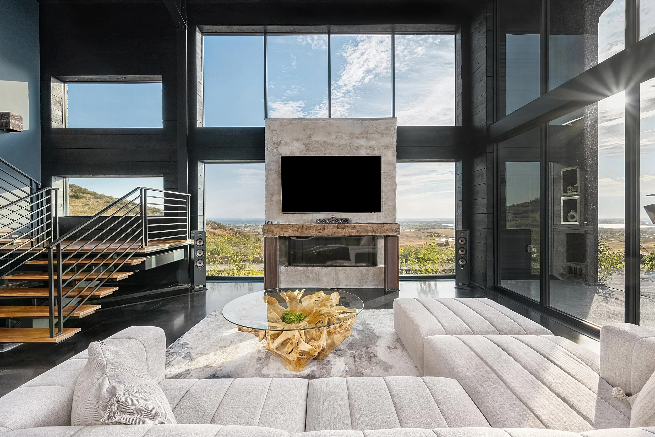 Modern living room with large windows, sectional sofa, glass table, and flat-screen TV.
