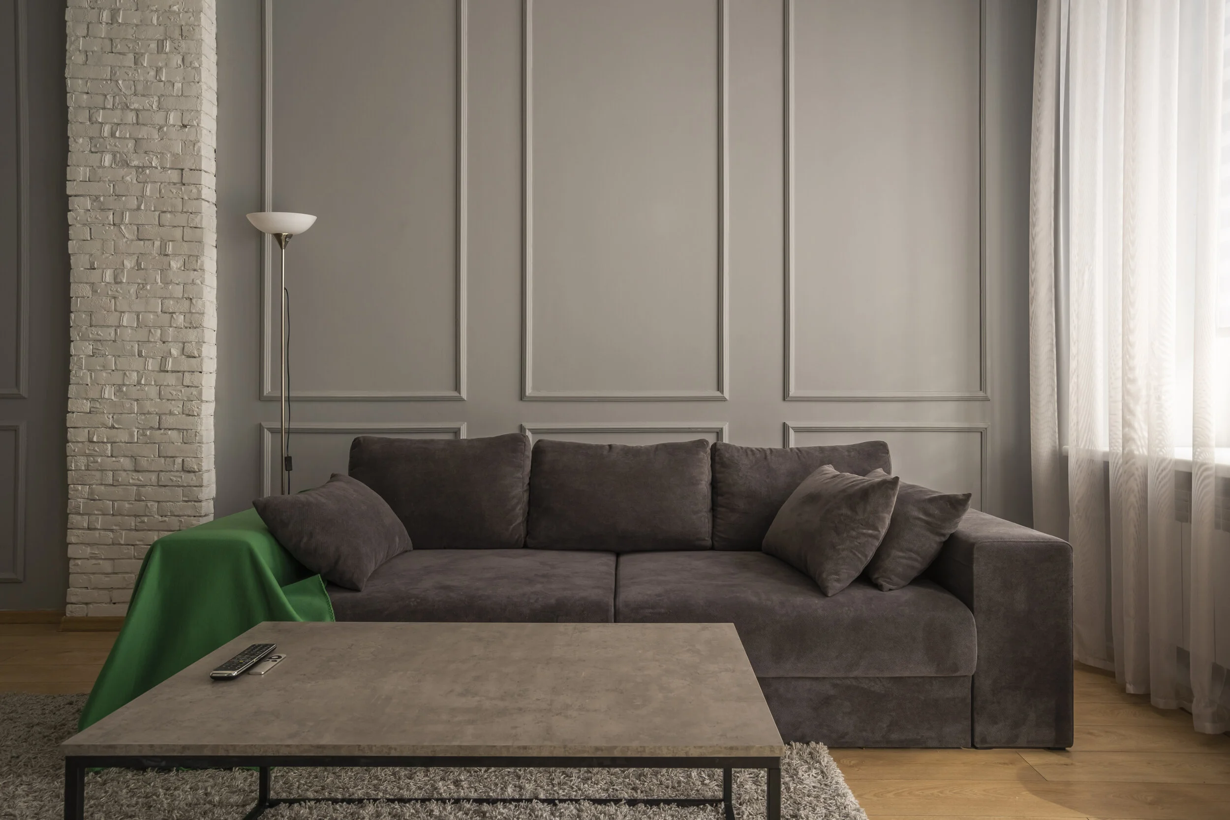 Living room with gray sectional sofa, green throw blanket, gray paneled wall, white brick pillar, tall floor lamp, and coffee table with remote control.