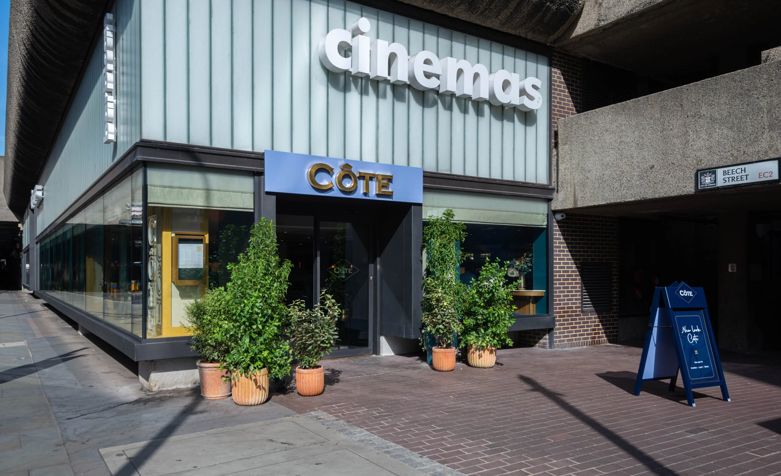 Facade of a restaurant named Côté with potted plants and an outdoor sign on a brick sidewalk, located on Beech Street, next to a cinema.