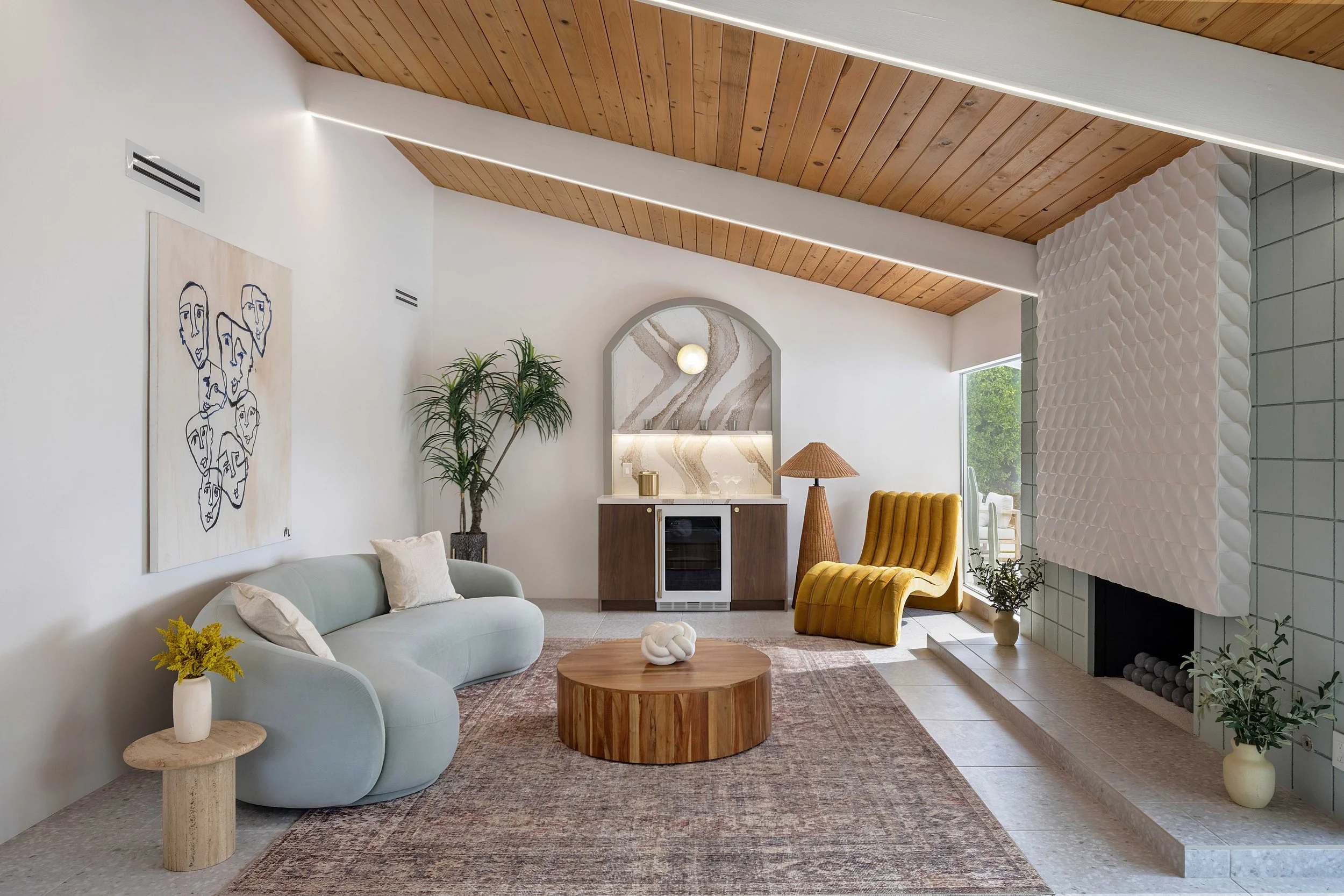 Mid-century modern living room with a light blue curved sofa, yellow lounge chair, wooden coffee table, potted plants, minimalist art, and a textured fireplace.