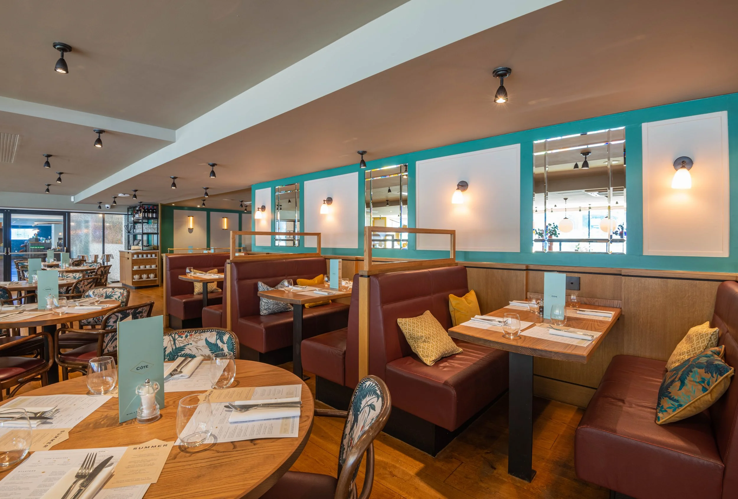 Interior of a modern restaurant with wooden tables, red leather booths, and decorative pillows. The space features teal accents, mirrors, and soft lighting. Tables are set with menus, glassware, and cutlery.