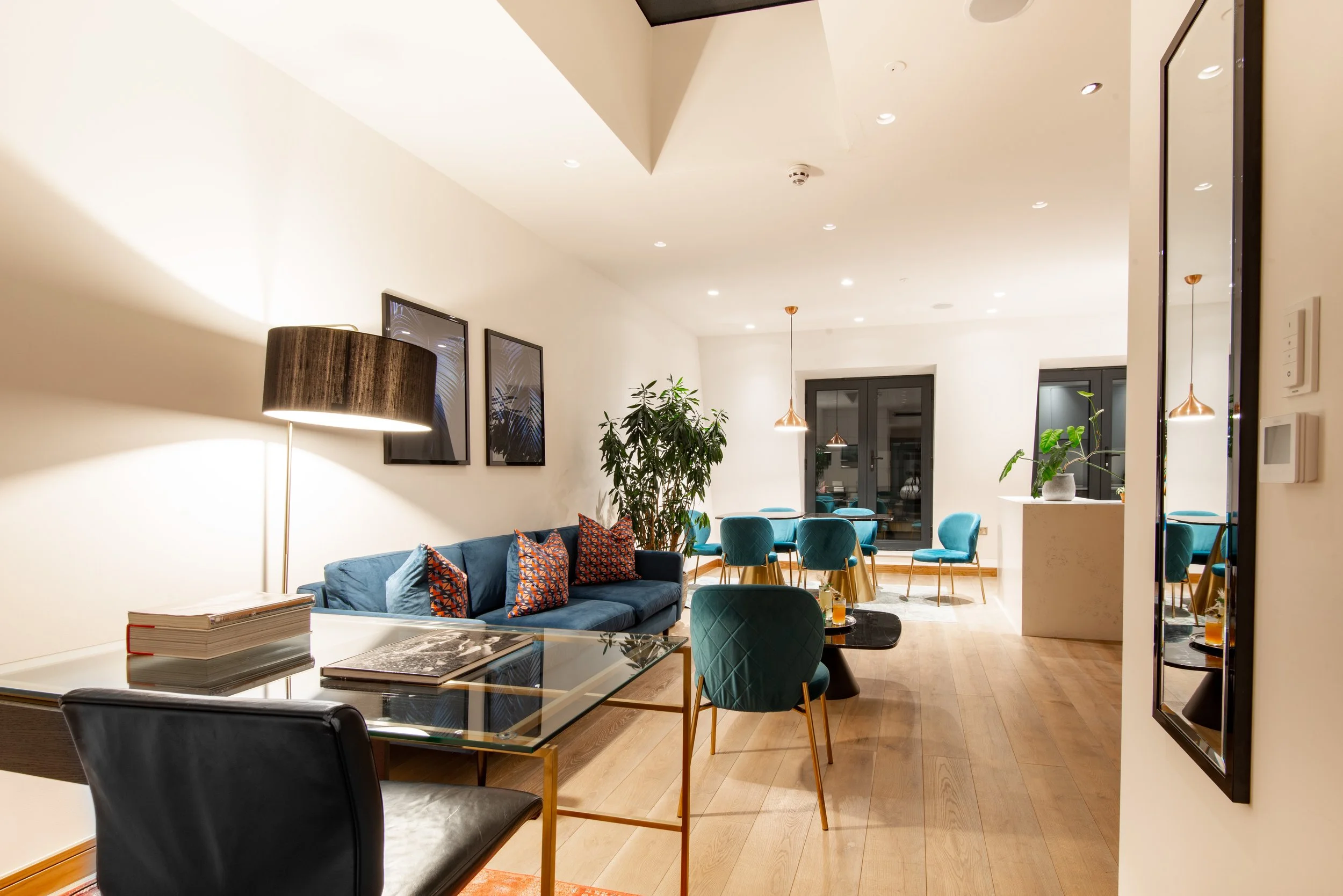 Modern living room with blue sofa, patterned cushions, glass table, and dining area with teal chairs and pendant lights.