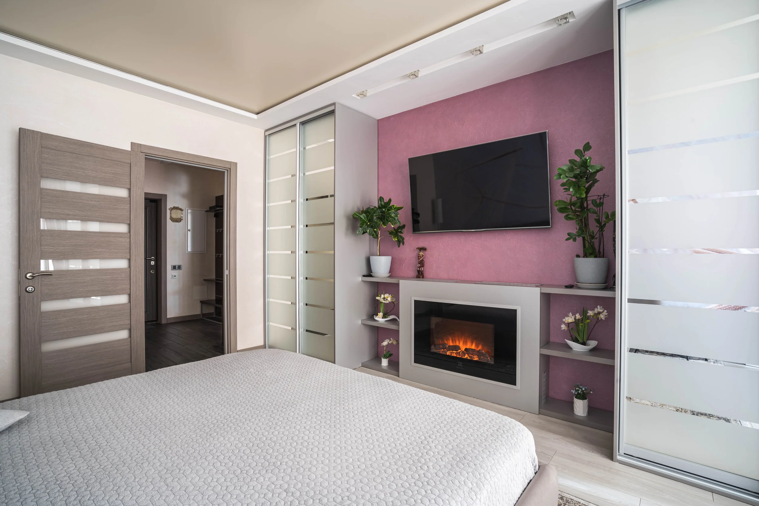 Modern bedroom interior with a bed, wall-mounted TV, and electric fireplace. The room features sliding closet doors, indoor plants, and a purple accent wall.