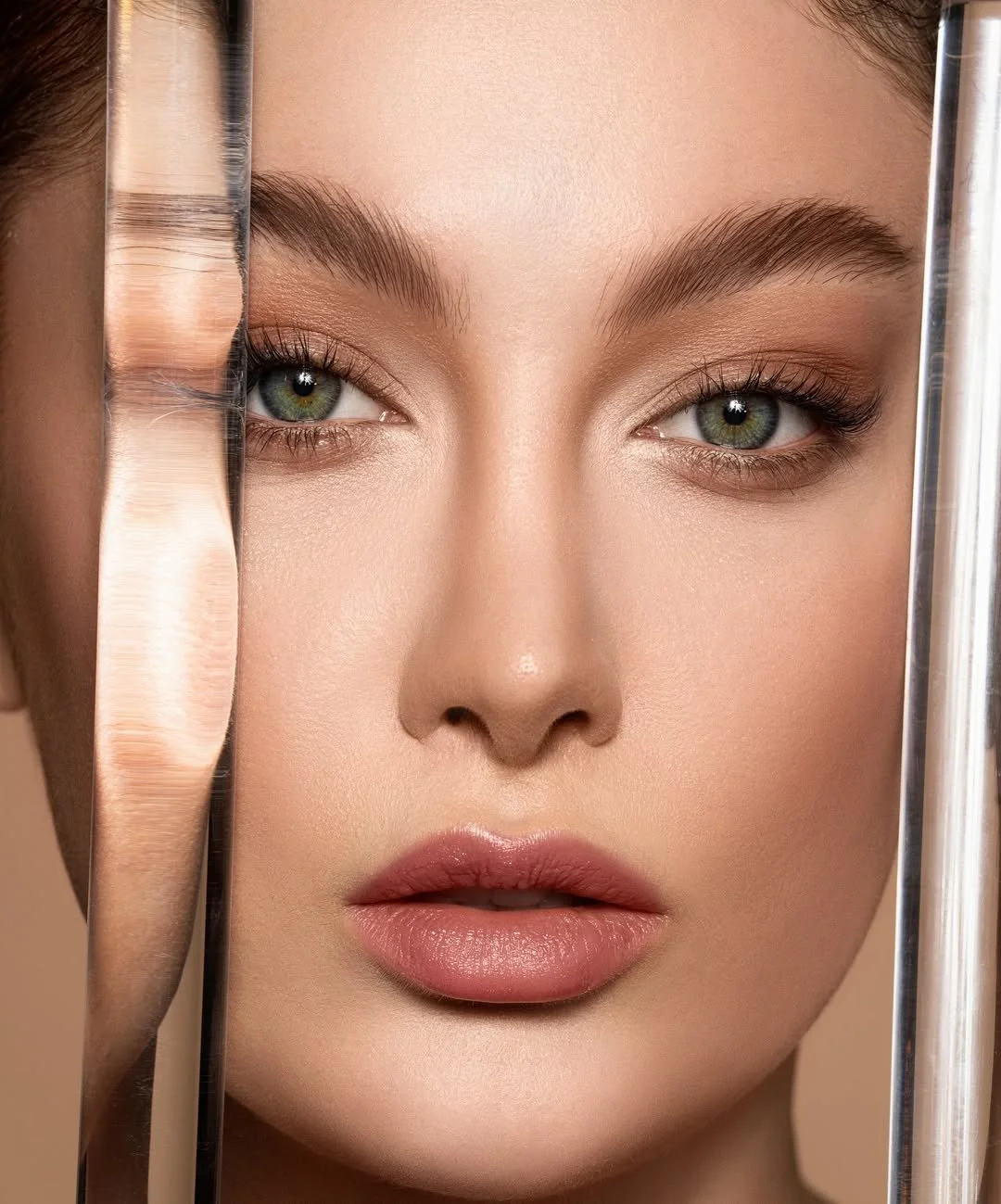 Close-up of a woman's face with clear skin, green eyes, and pink lips, framed by blurred glass objects.