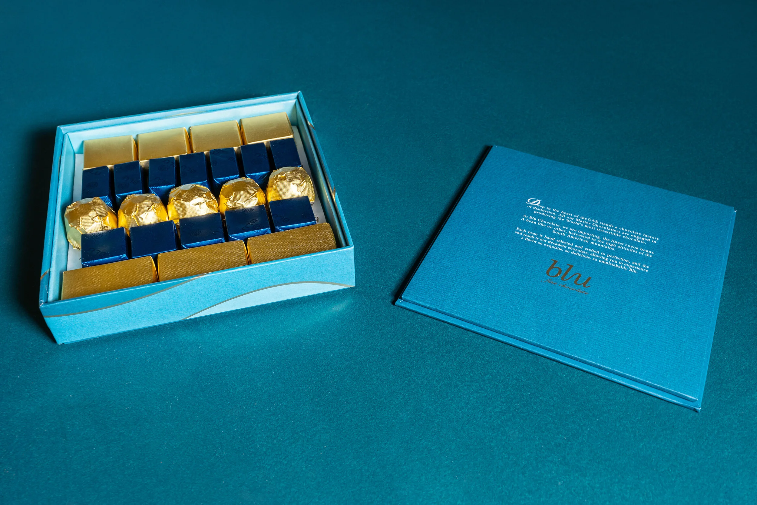 Box of blue and gold wrapped chocolates with a lid on a teal surface.