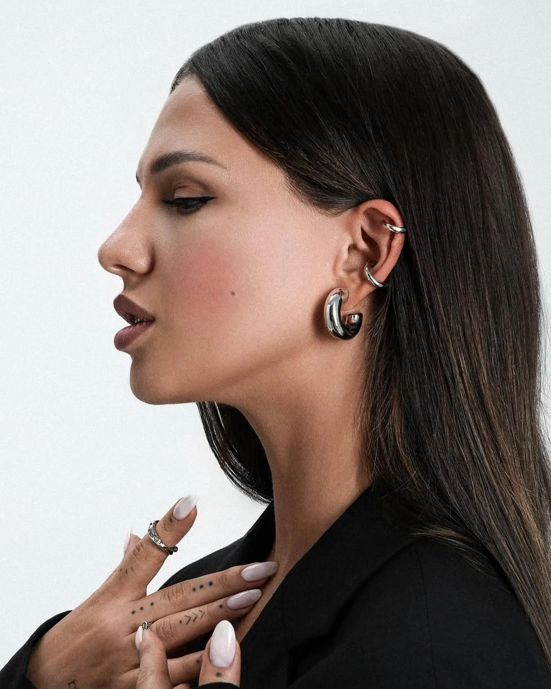 Profile of a woman wearing multiple silver earrings and rings, with tattoos on her fingers and long dark hair against a white background.