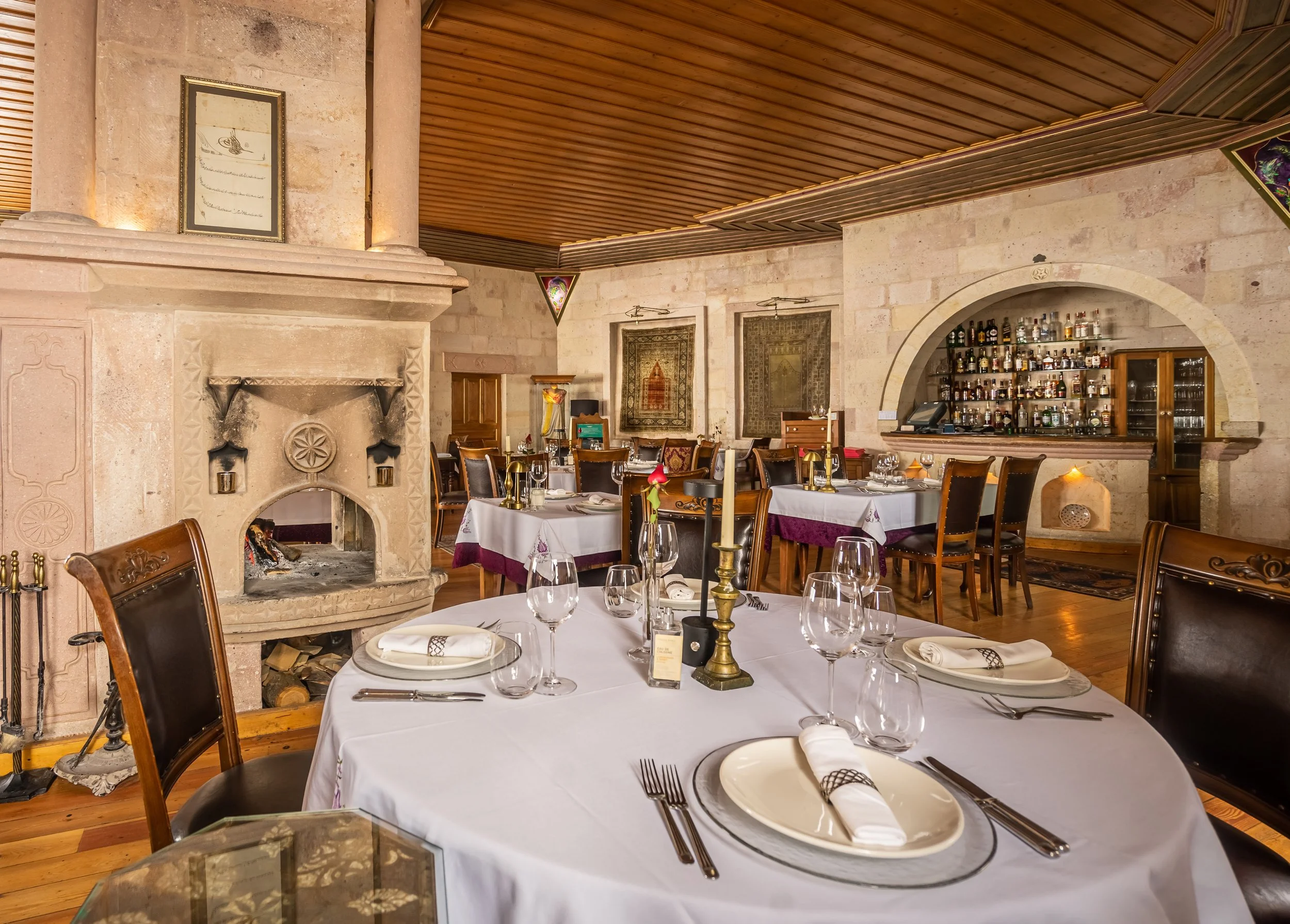 Elegant restaurant interior with wooden ceiling, stone fireplace, round tables set with white cloths, plates, and wine glasses, and a bar stocked with various bottles.