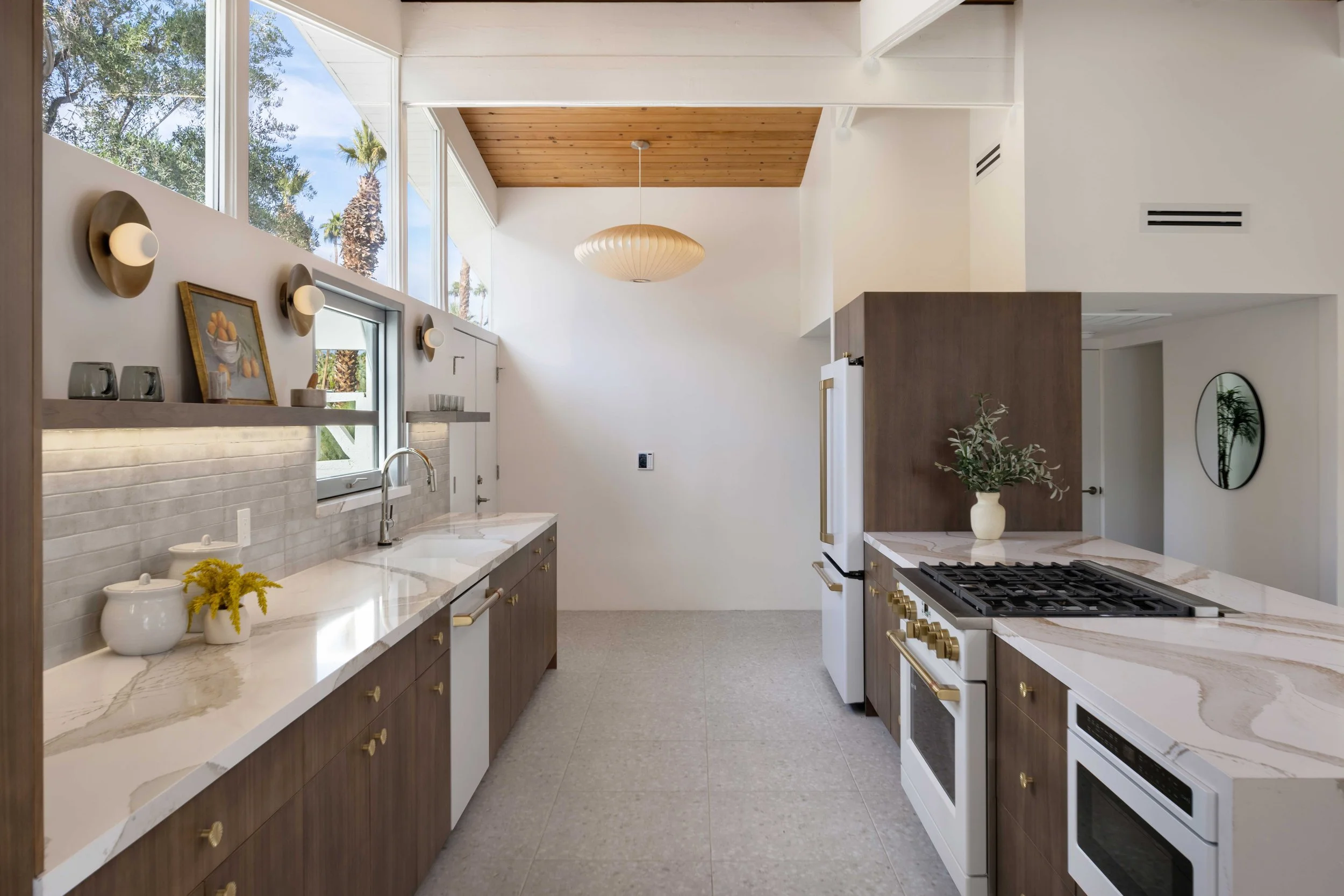 Modern kitchen with wooden cabinets, white countertops, built-in stovetop and oven, large windows, pendant light, and decorative plants.