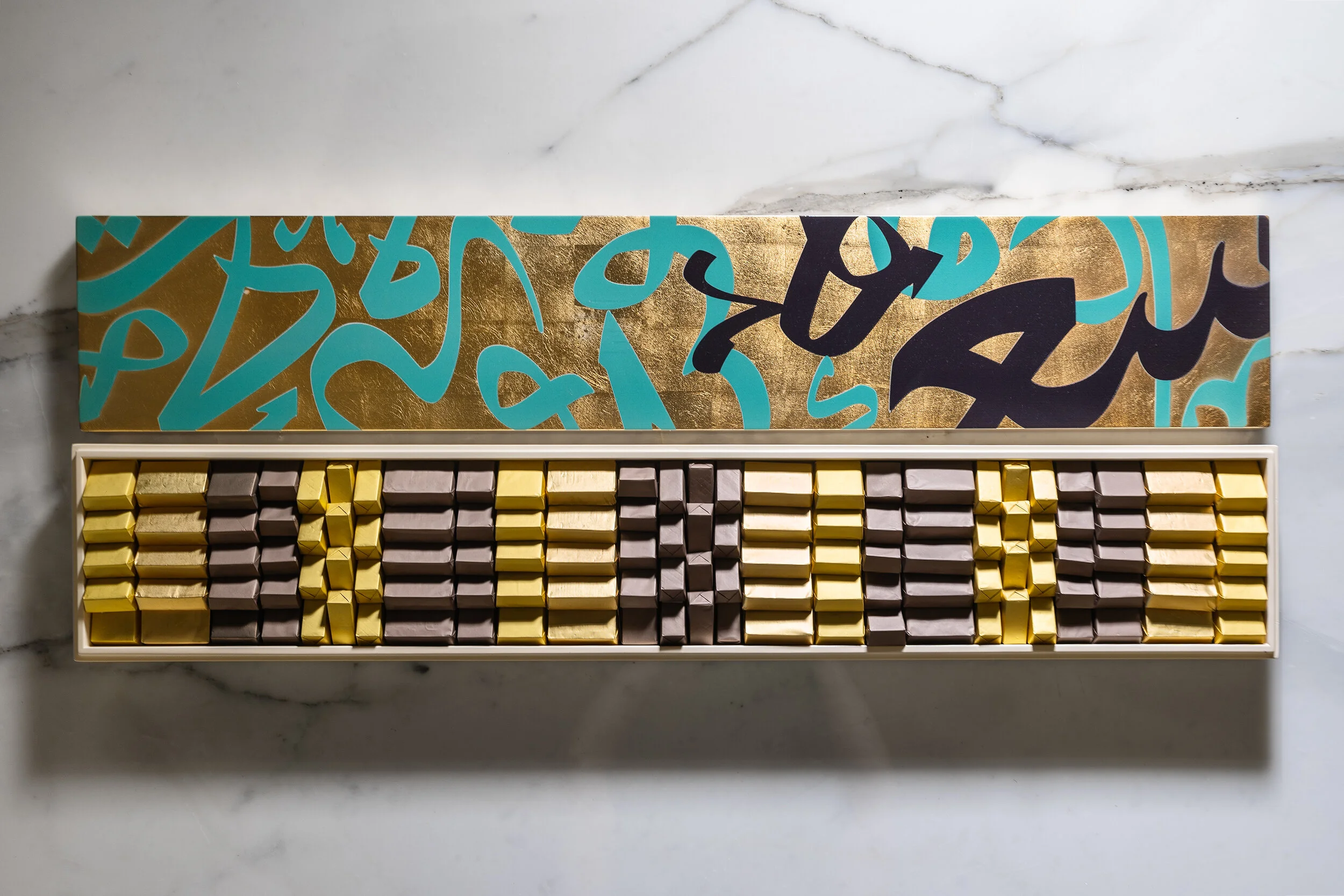 Open box of chocolates arranged neatly with gold and brown wrappers, featuring a decorative, colorful lid design on a marble surface.