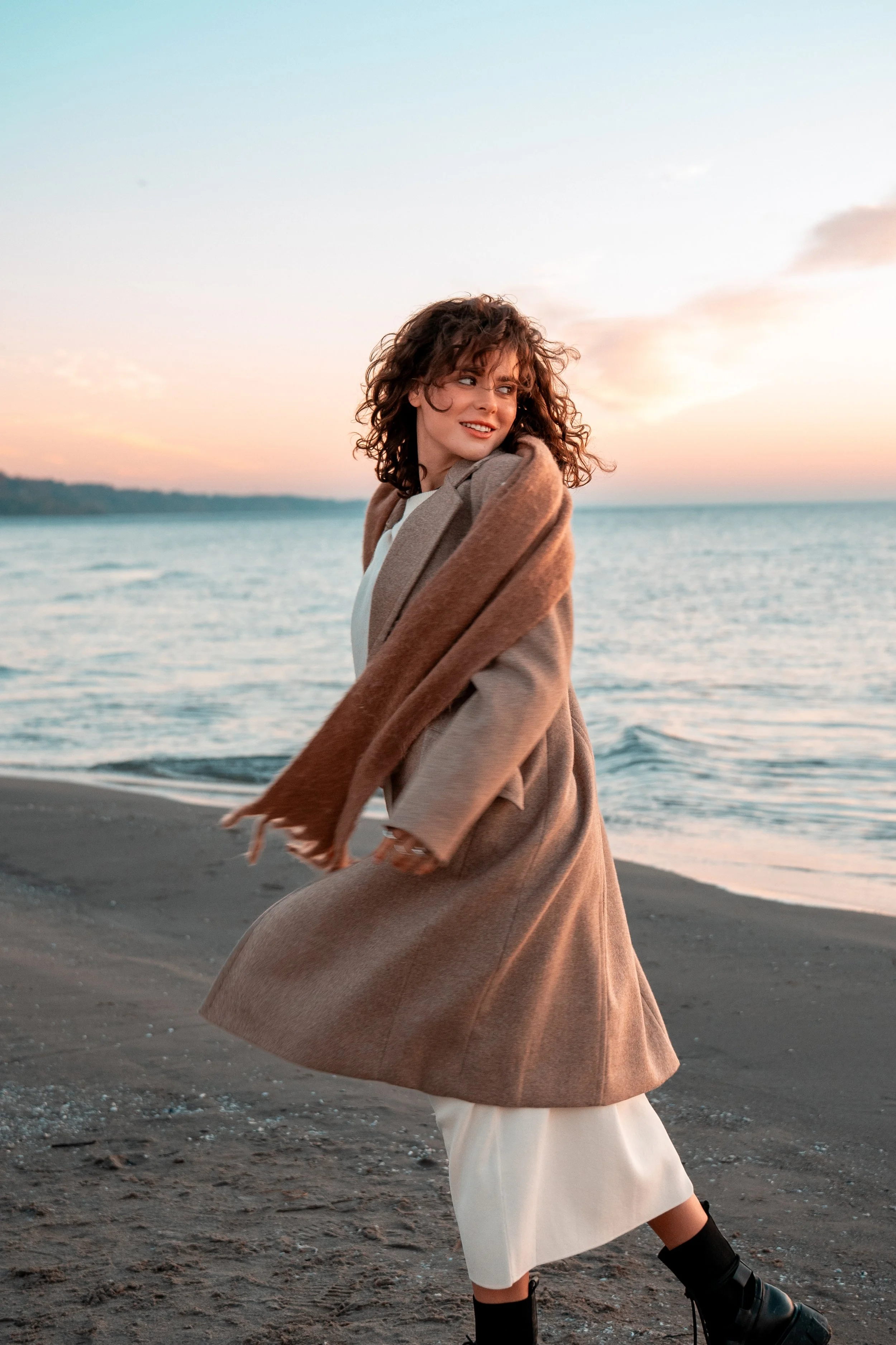 Woman in coat and scarf walking on beach at sunset