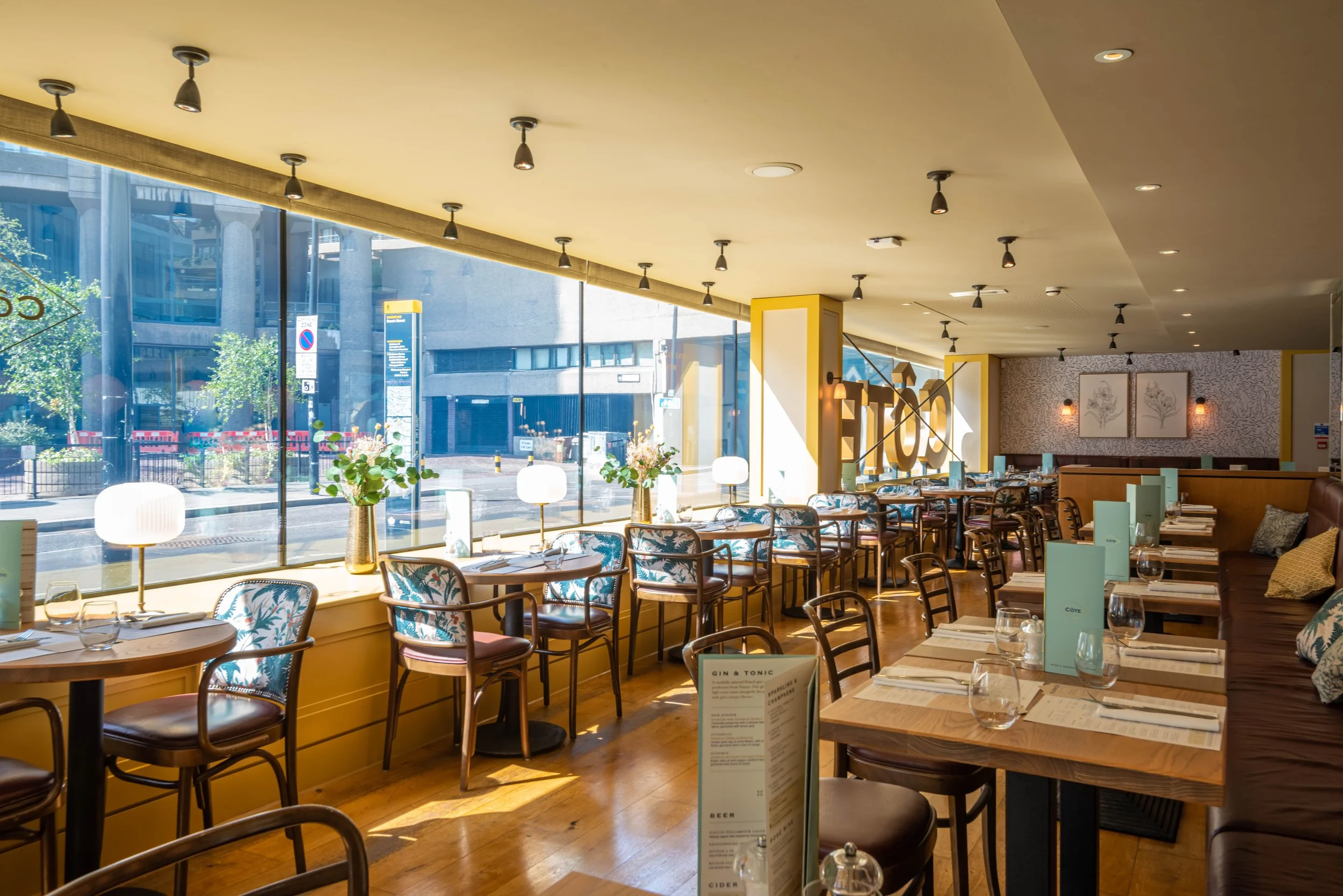 Interior of a modern restaurant with wooden tables and chairs, decorative cushions, and large windows offering a street view. Elegant table settings include menus, glassware, and vases with flowers. Natural light fills the room.