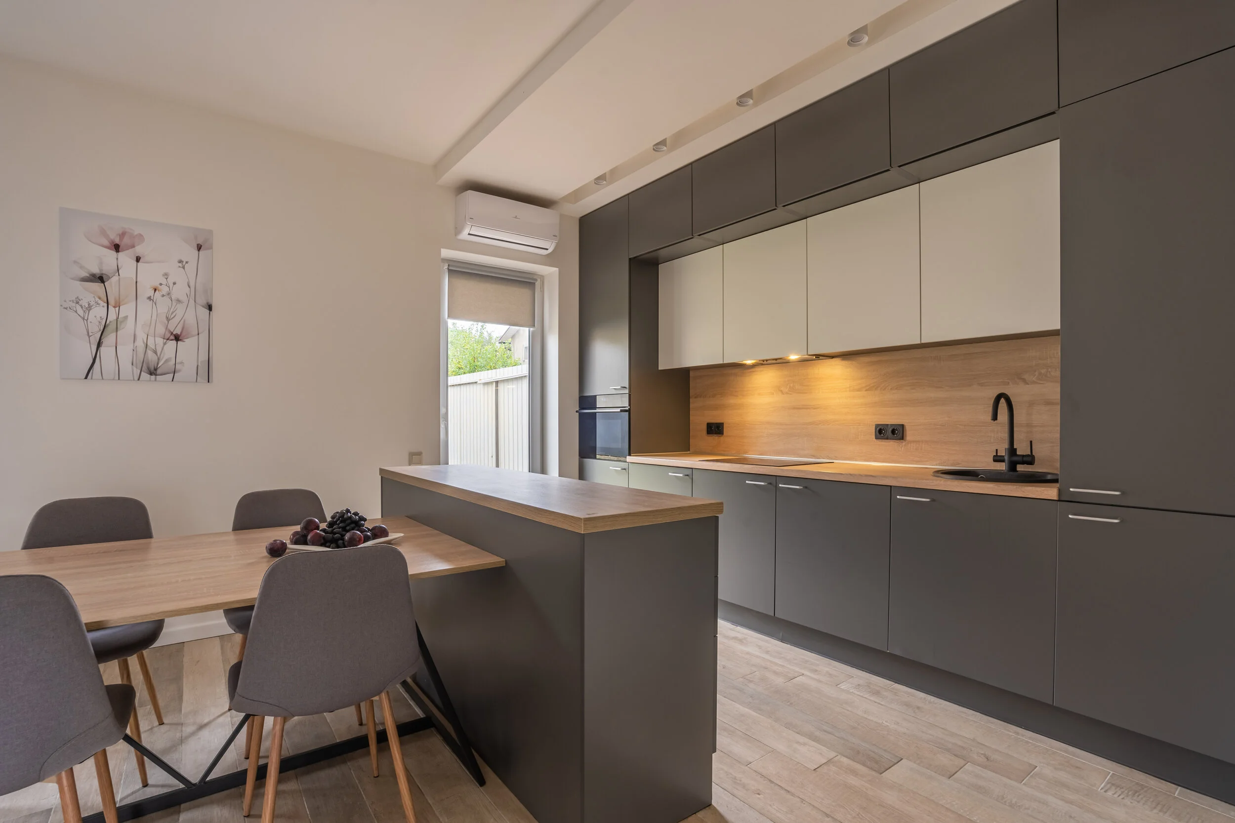 Modern kitchen with gray cabinets, wooden countertops, integrated appliances, a dining table with chairs, and a wall-mounted floral artwork.