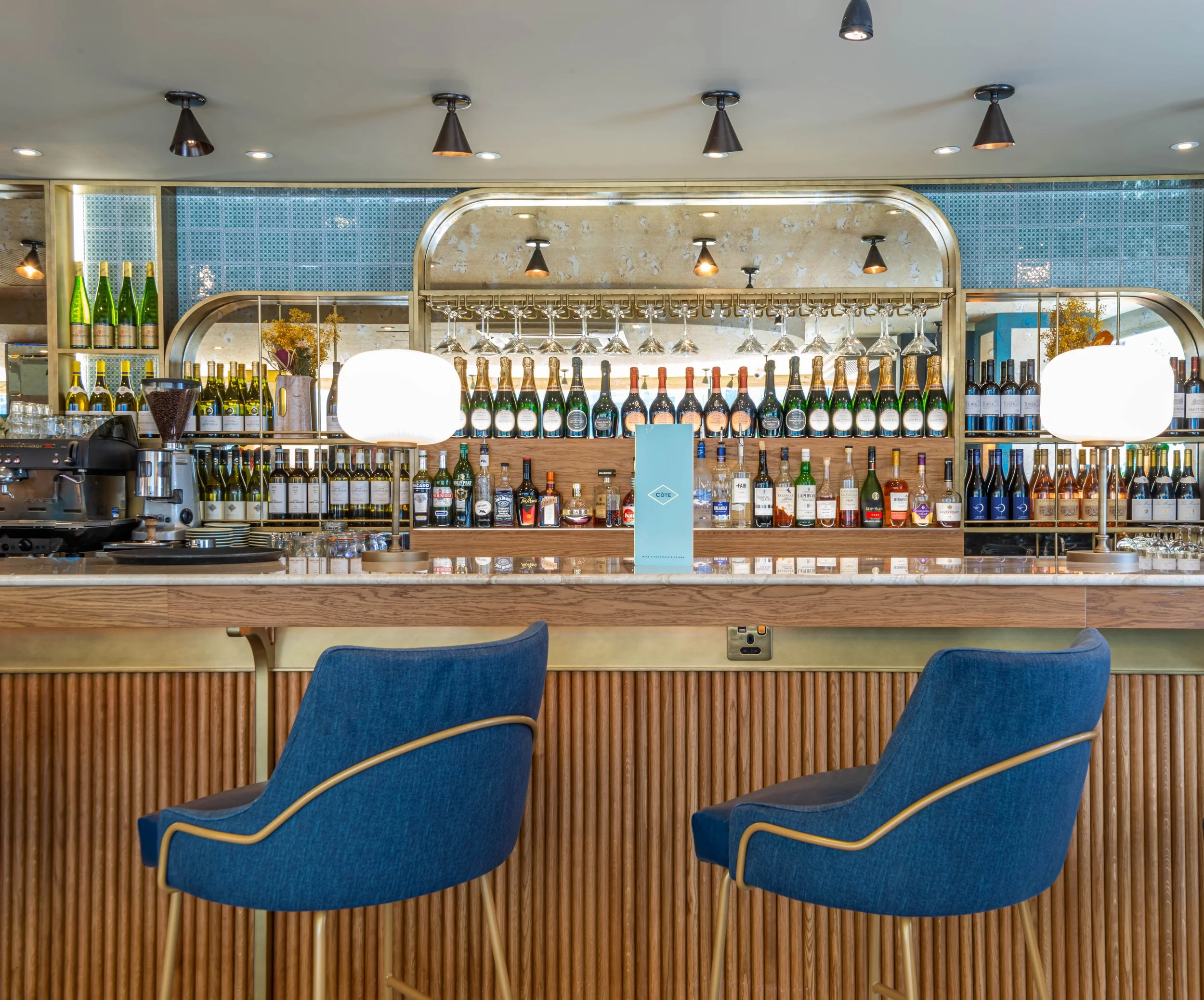 Modern bar interior with blue chairs and a wooden counter, featuring a well-stocked selection of wine and liquor bottles on shelves. Overhead lights illuminate the bar area.