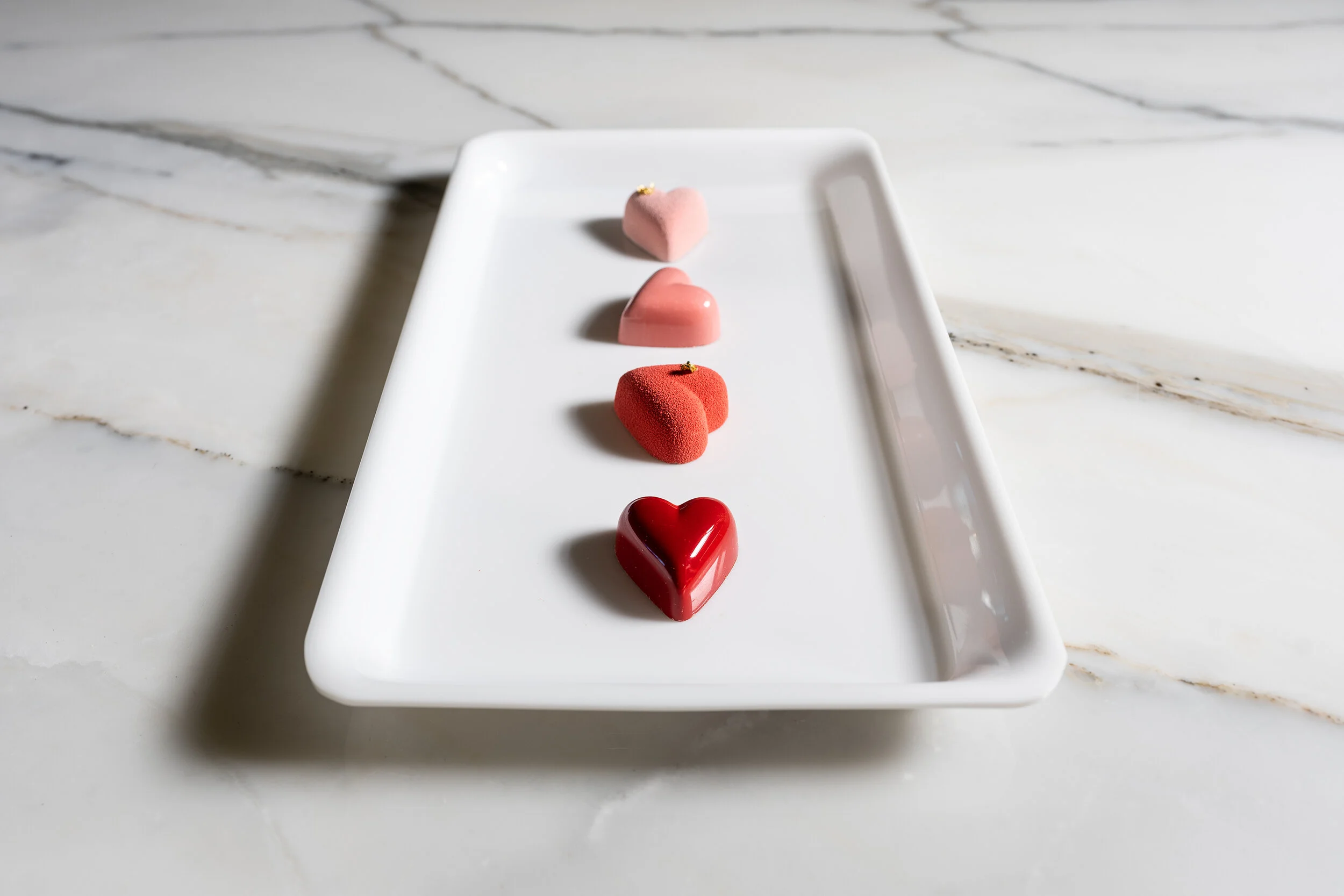 Four heart-shaped desserts on a rectangular white plate on a marble surface.