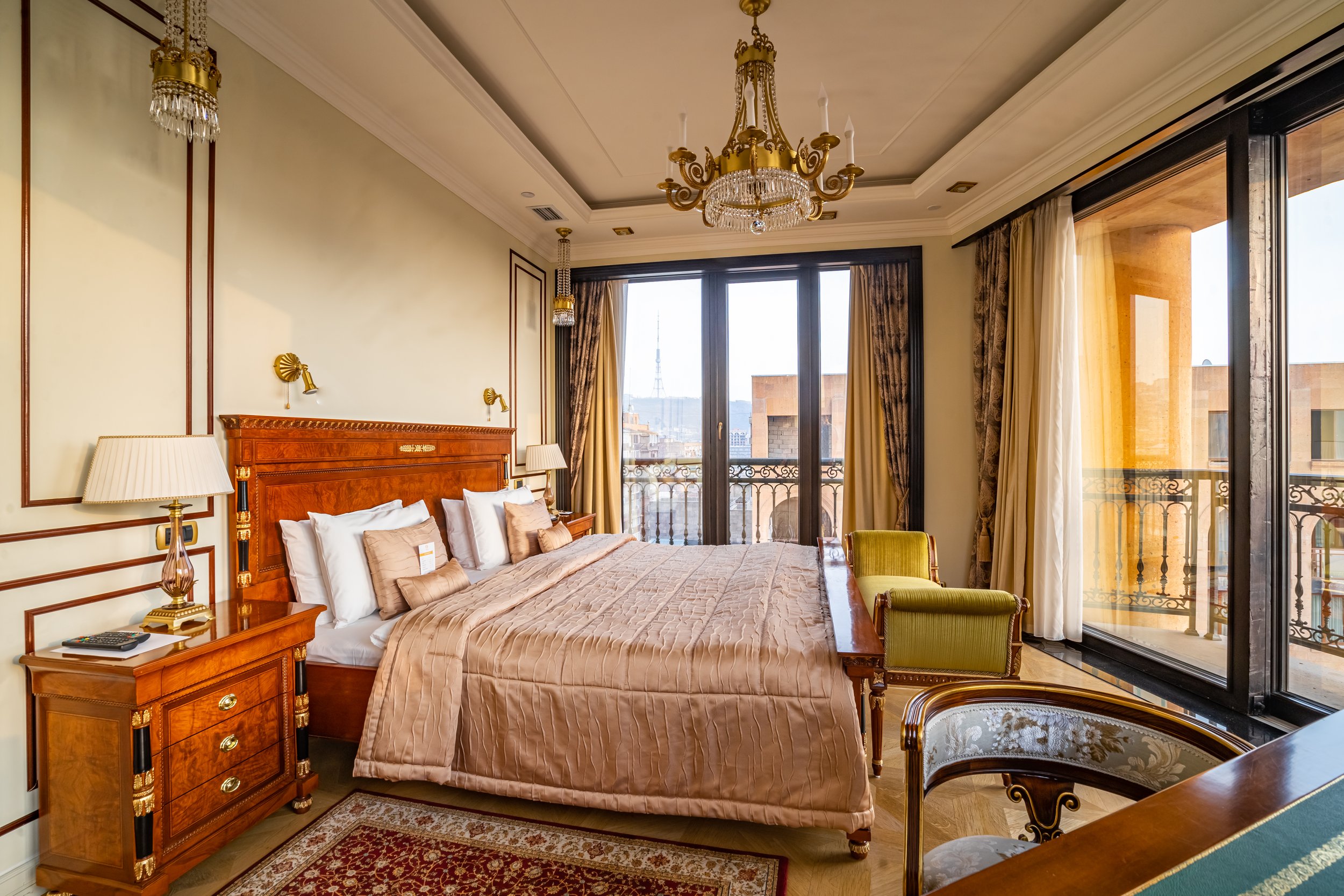 Elegant bedroom with a large bed, ornate wooden furniture, and a chandelier. The room has large windows with a view, beige and gold color scheme, and rich-textured fabrics.