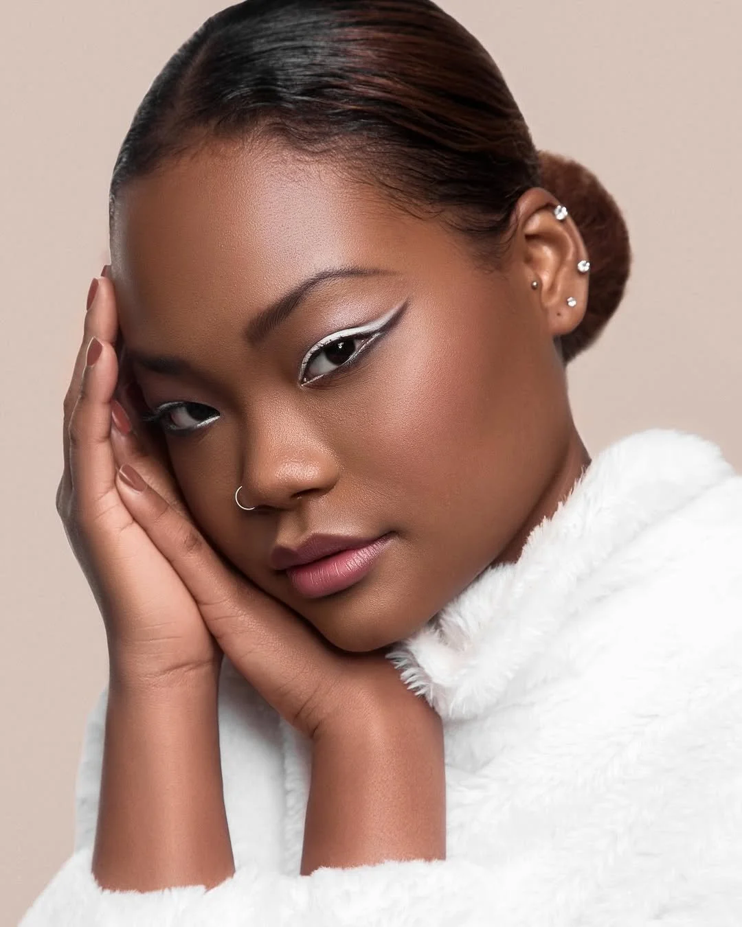 Close-up of a person with intricate eye makeup, wearing a white garment, resting their face on their hands. They have a nose ring and multiple ear piercings.