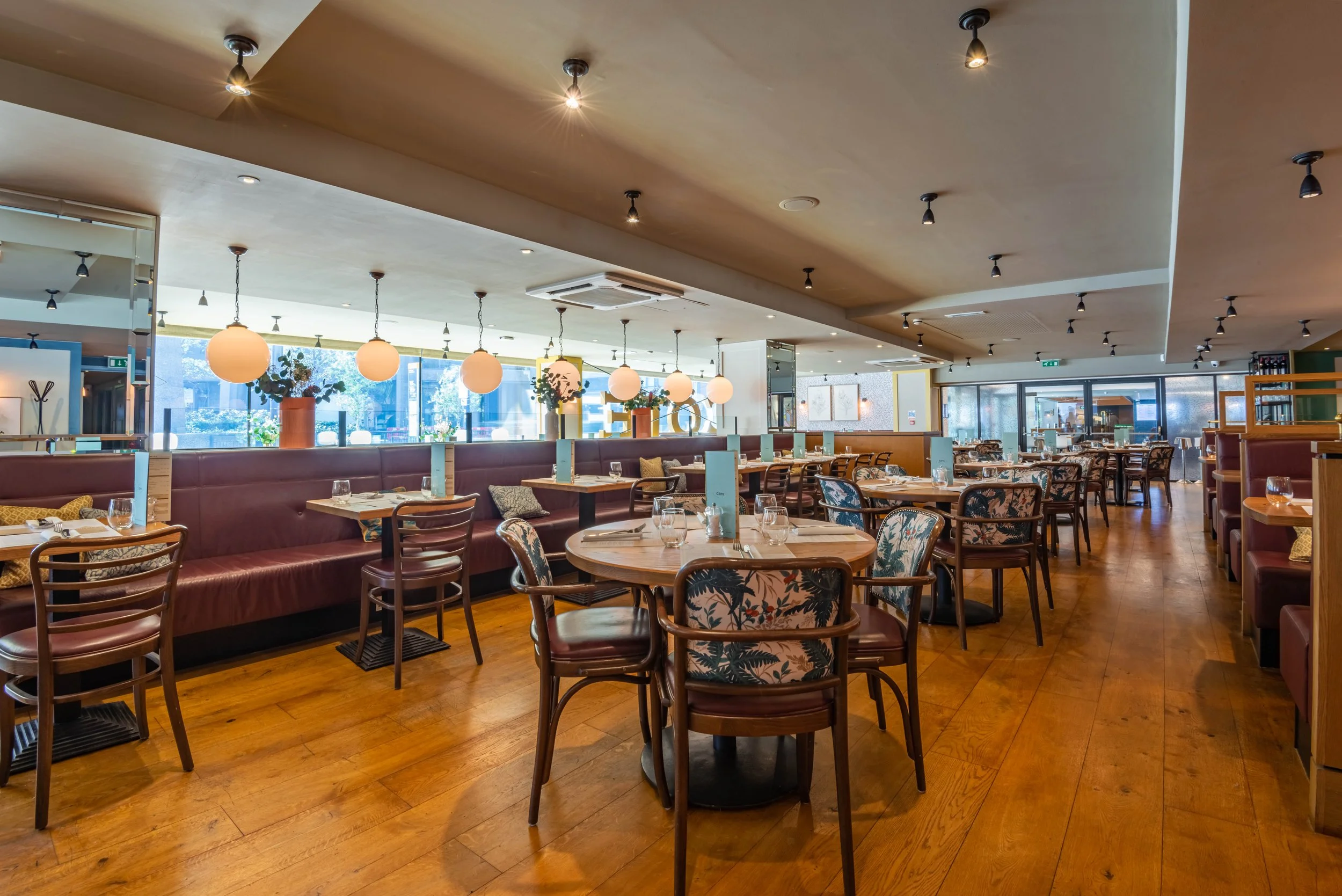 Interior of a modern, spacious restaurant with wooden floors, round tables, cushioned chairs, and warm lighting.