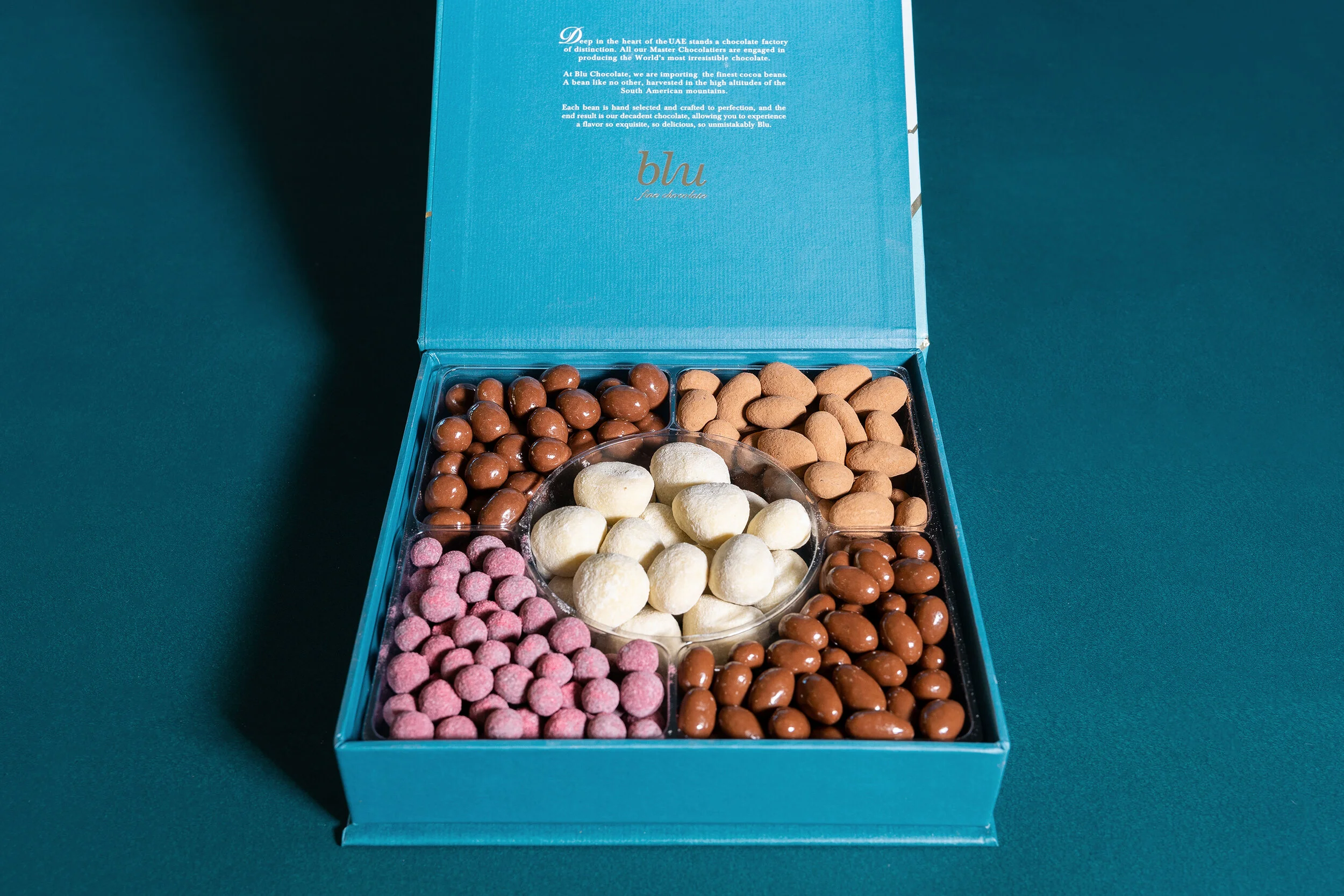 An open box of assorted chocolates and nuts, including chocolate-covered nuts, pink sugar-coated candies, white powdered treats, and plain nuts.