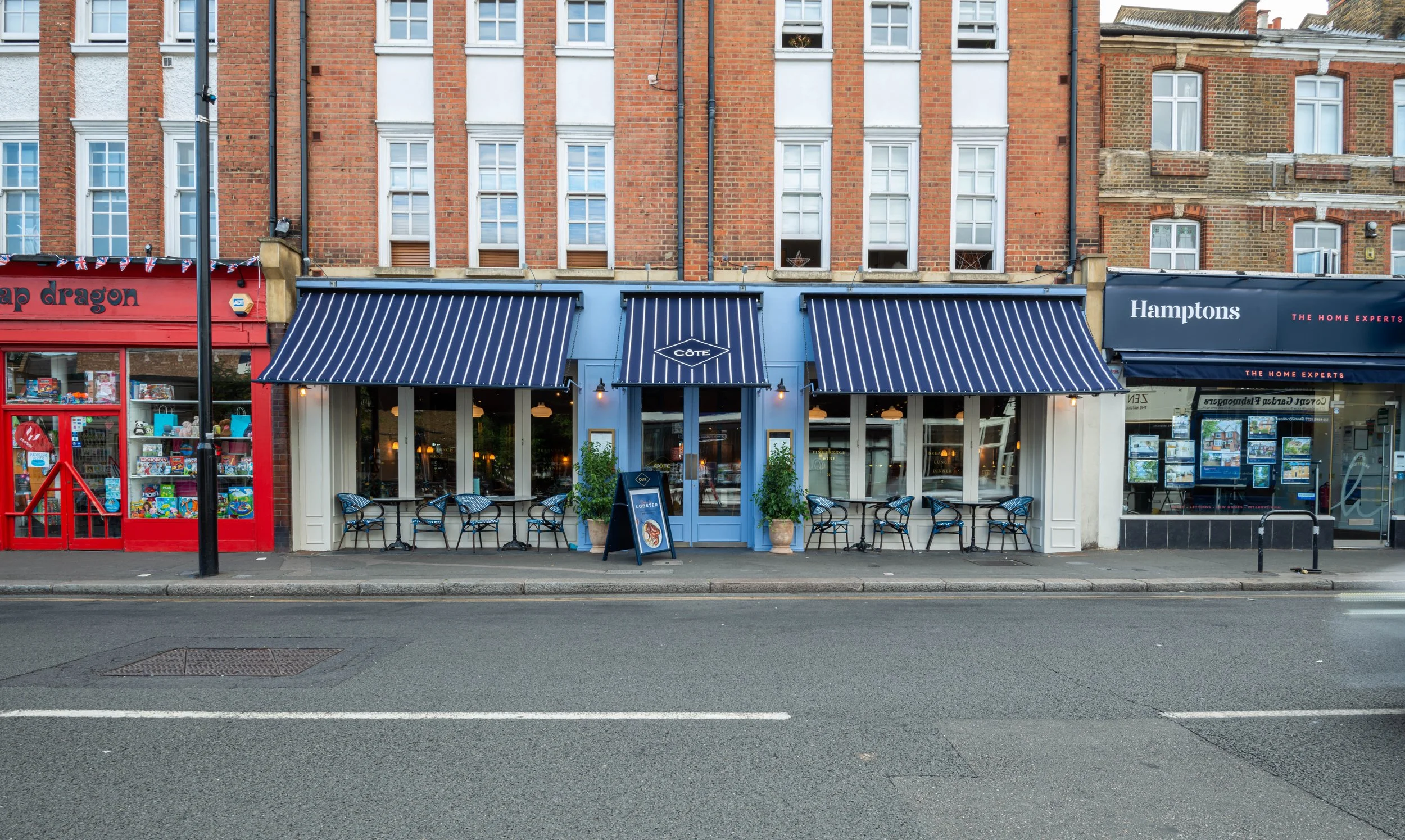 Street view of a row of shops, including a toy store with a red facade, a cafe with blue striped awnings, and a real estate office with a dark blue sign. Outdoor seating is visible at the cafe.