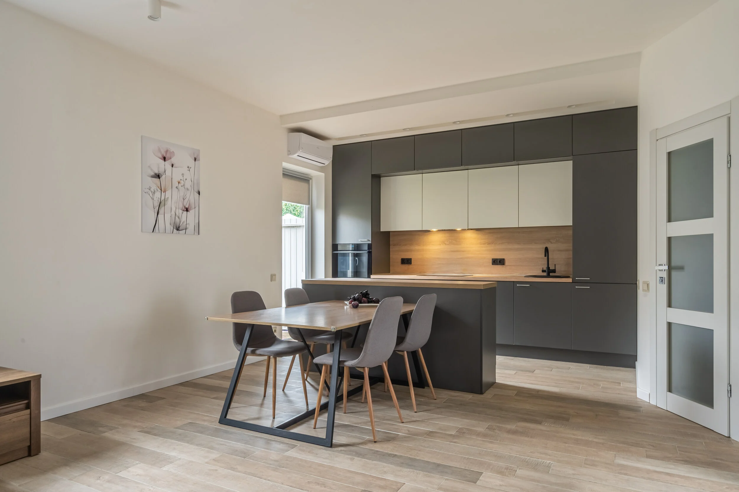 Modern kitchen with dining area, featuring gray cabinets, wooden countertop, dining table with chairs, wood flooring, and a wall art of flowers.