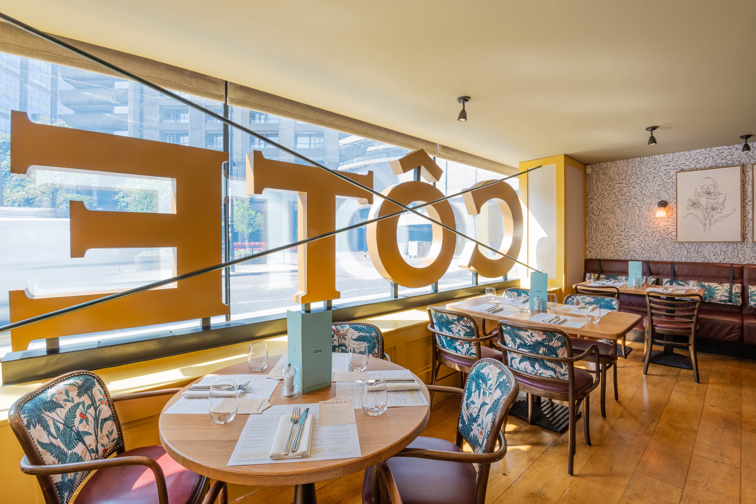 Interior of a cozy café with round wooden tables, patterned chairs, and a large window displaying the word 'CÔTE'. The room is decorated with wallpaper, framed artwork, and ambient lighting.