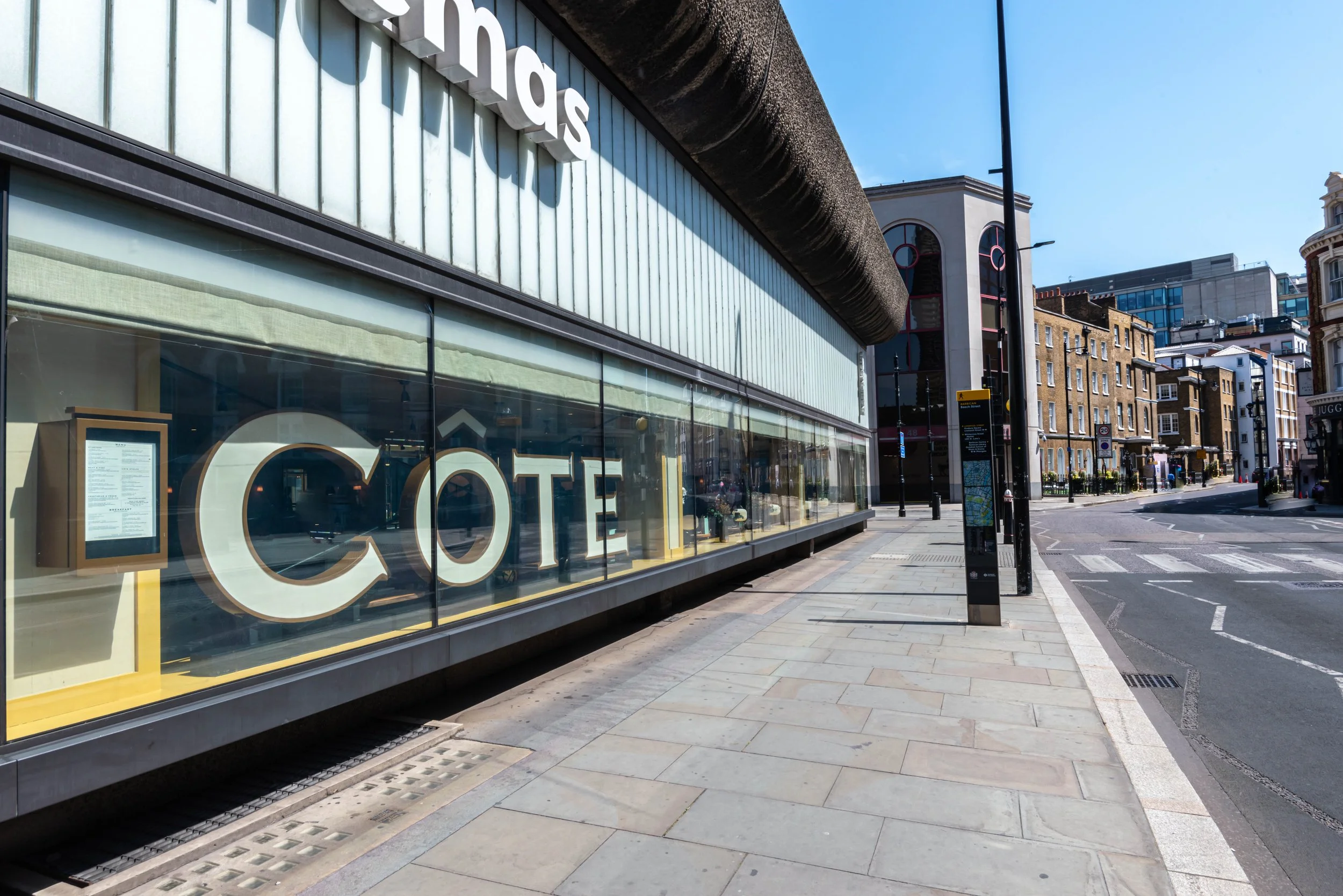Street view of a building with large glass windows displaying the word "CÔTE," adjacent to a quiet, urban street.