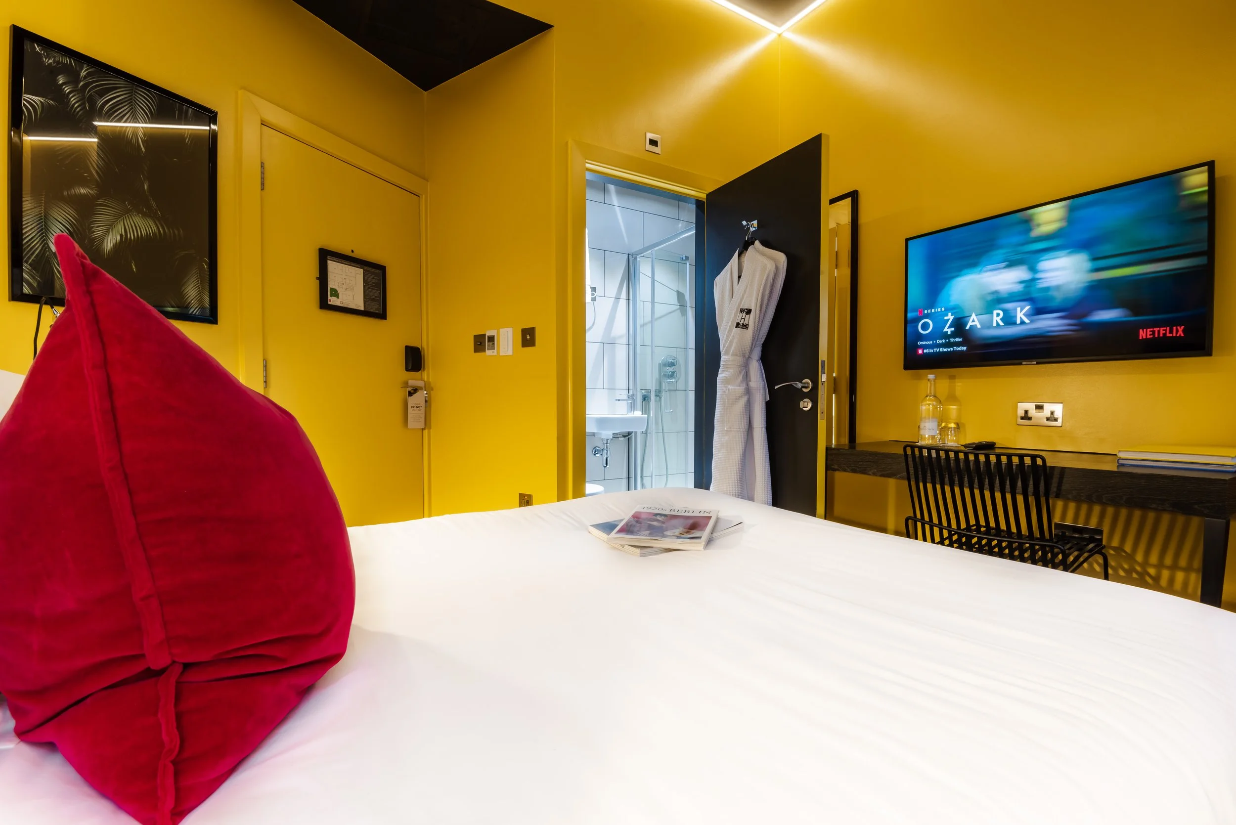 Hotel room with yellow walls, a bed with a red pillow, a wall-mounted TV displaying a Netflix show, a black chair and desk, bathroom visible, and a white robe hanging on the door.