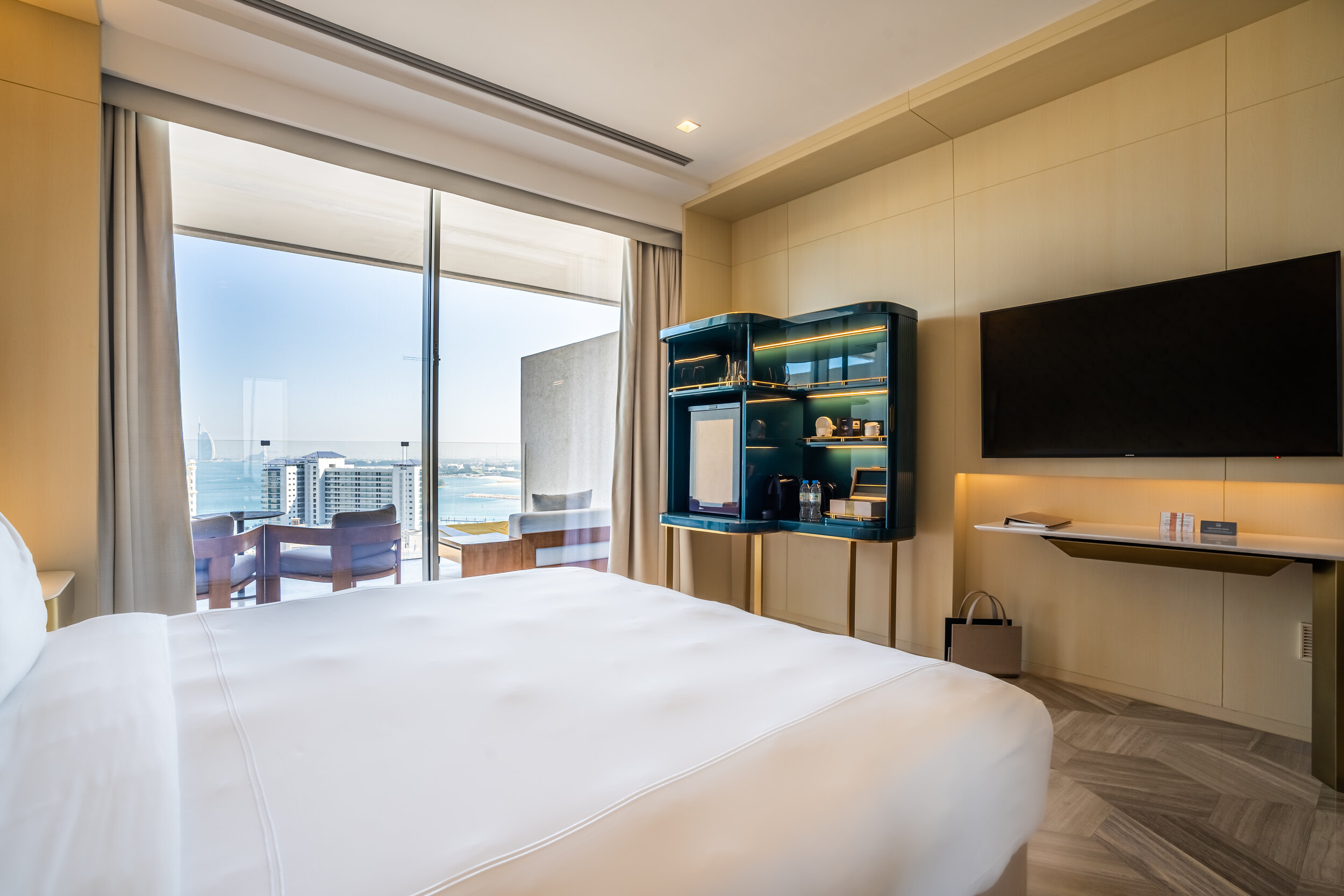 Modern hotel room with large bed, wall-mounted TV, minibar, and a view of buildings and water through a sliding glass door.