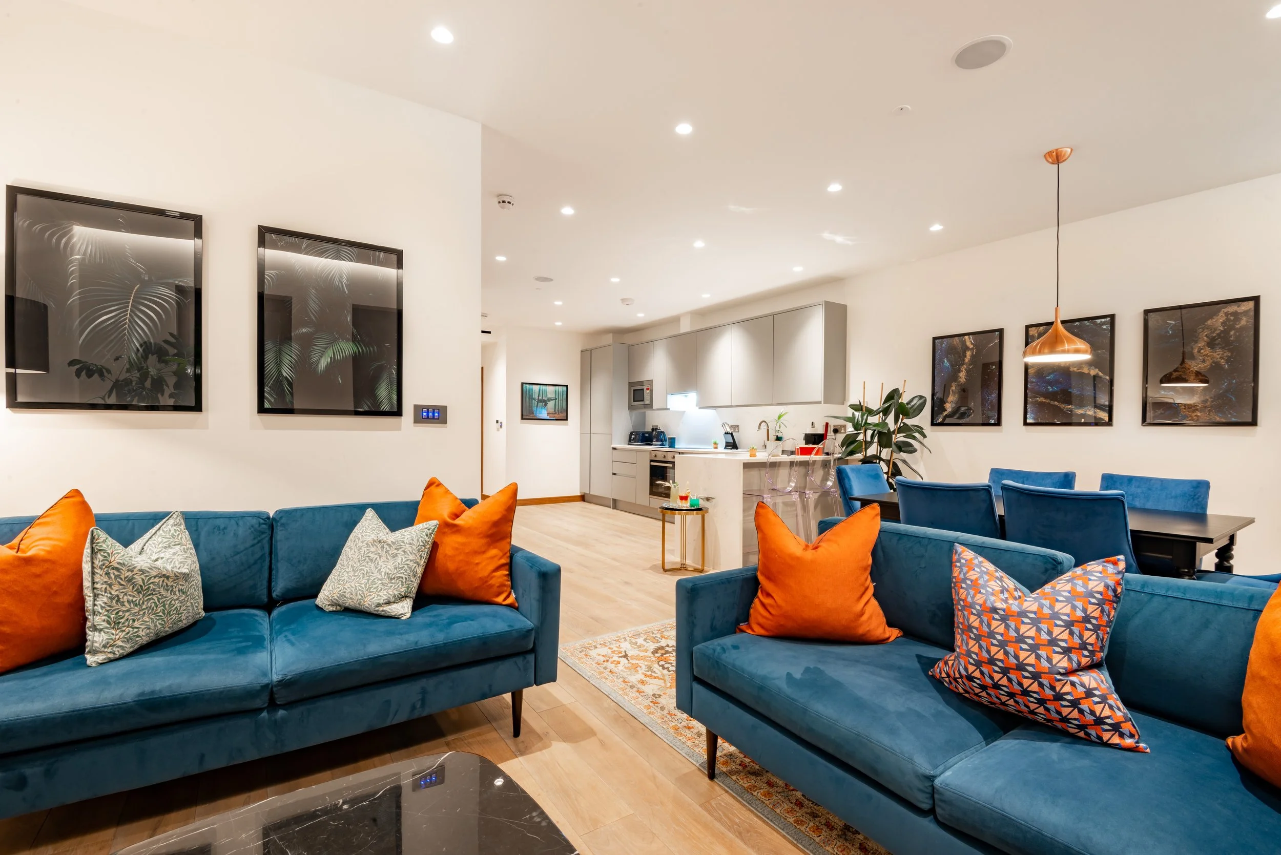 Modern open-concept living area with blue sofas, orange and patterned cushions, a dining table, kitchen with grey cabinets, and framed artwork on the walls.