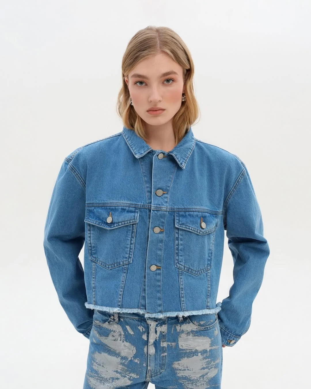 Person wearing a blue denim jacket with button details and ripped denim jeans, standing against a plain white background.