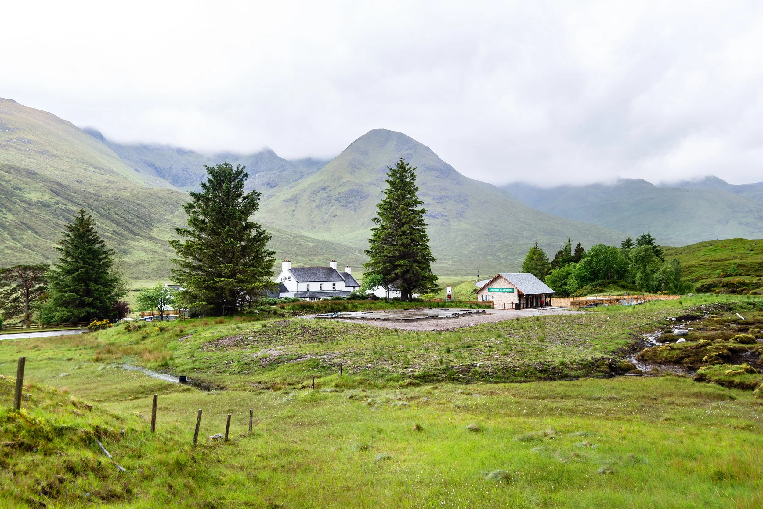 Scenic landscape of a grassy field with trees and two buildings, set against rolling green hills and misty mountains in the background.