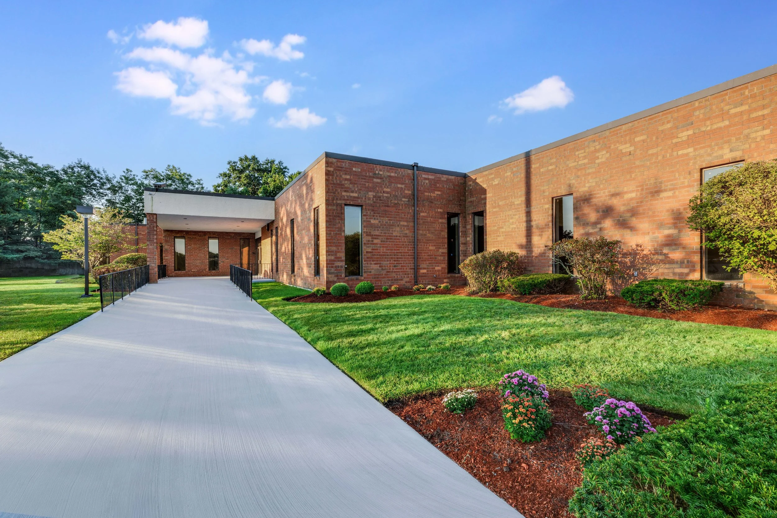 Modern brick office building with large windows, surrounded by a well-kept lawn, bushes, and flower beds, under a clear blue sky.