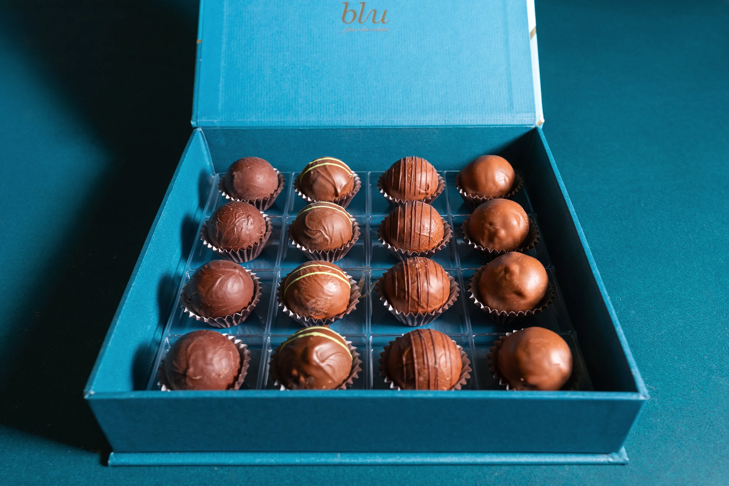 Open blue box with sixteen assorted chocolate truffles arranged in a grid.