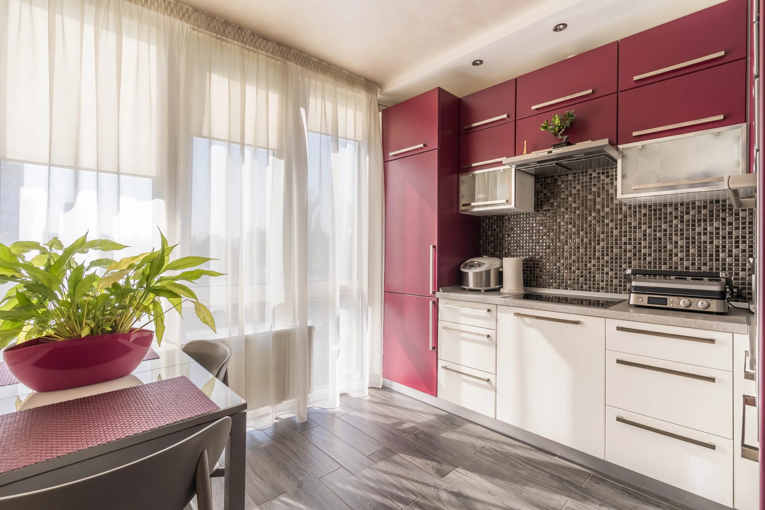 Modern kitchen with red cabinets, white countertops, mosaic backsplash, and stainless steel appliances, featuring a plant on the dining table with natural light streaming through sheer curtains.