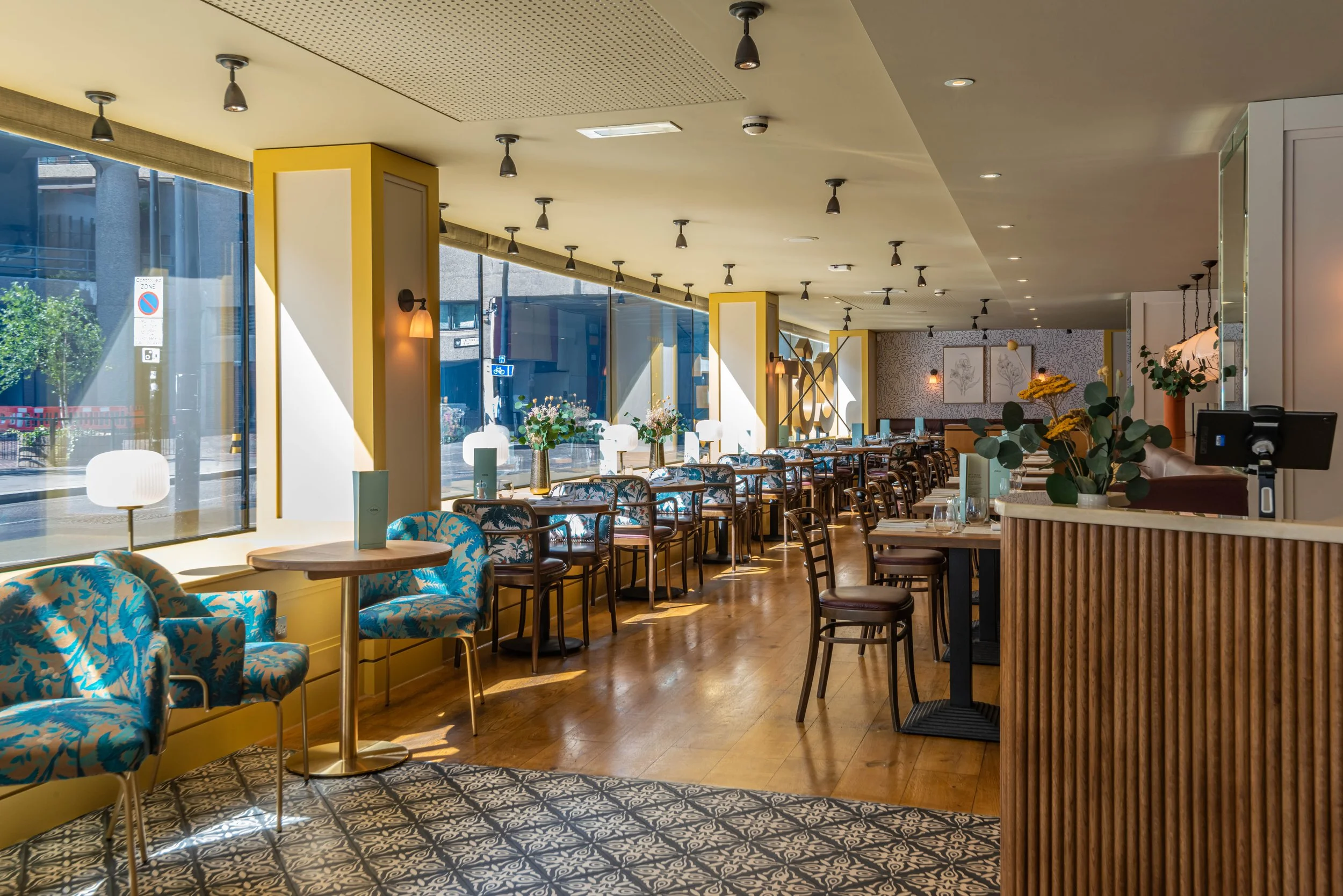 Modern, brightly lit restaurant interior with patterned chairs and tables arranged in rows, large windows, and decorative plants and lighting.