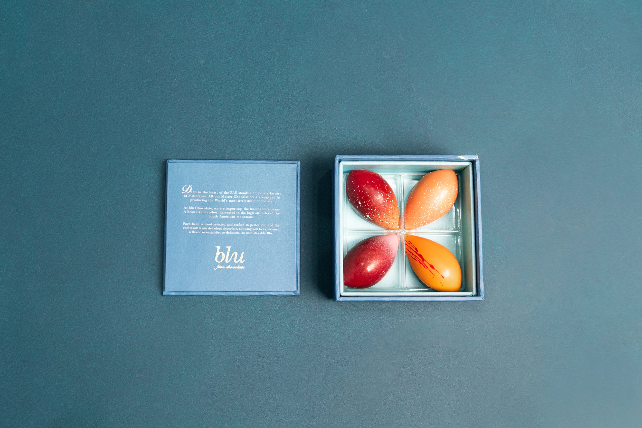 Open box of four artisan chocolates with textured, speckled designs and a closed box with text description on a blue surface.