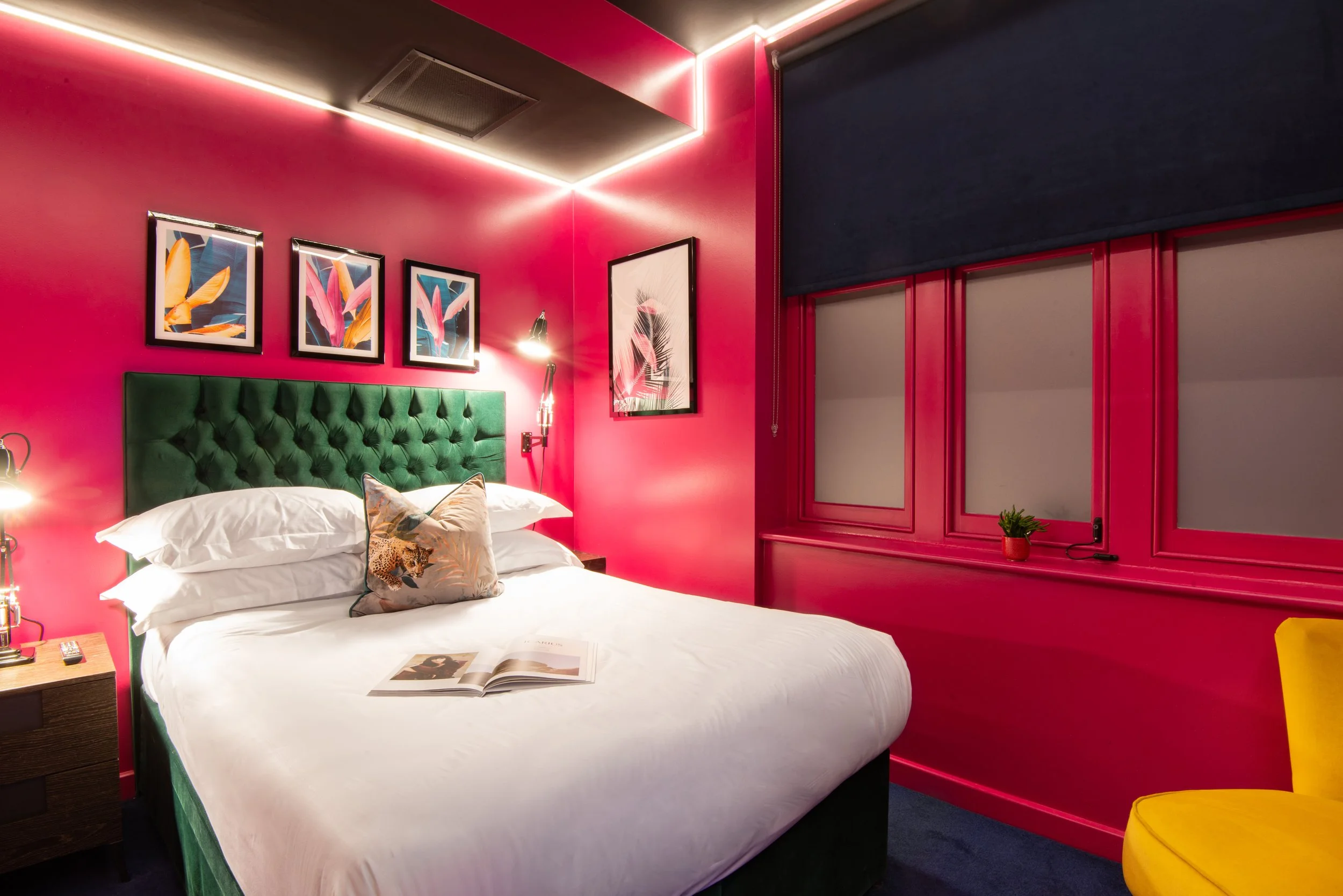 Colorful hotel room with pink walls, green tufted headboard, white bedding, and artwork above the bed. A small plant sits on the windowsill, and a yellow chair is visible in the corner.
