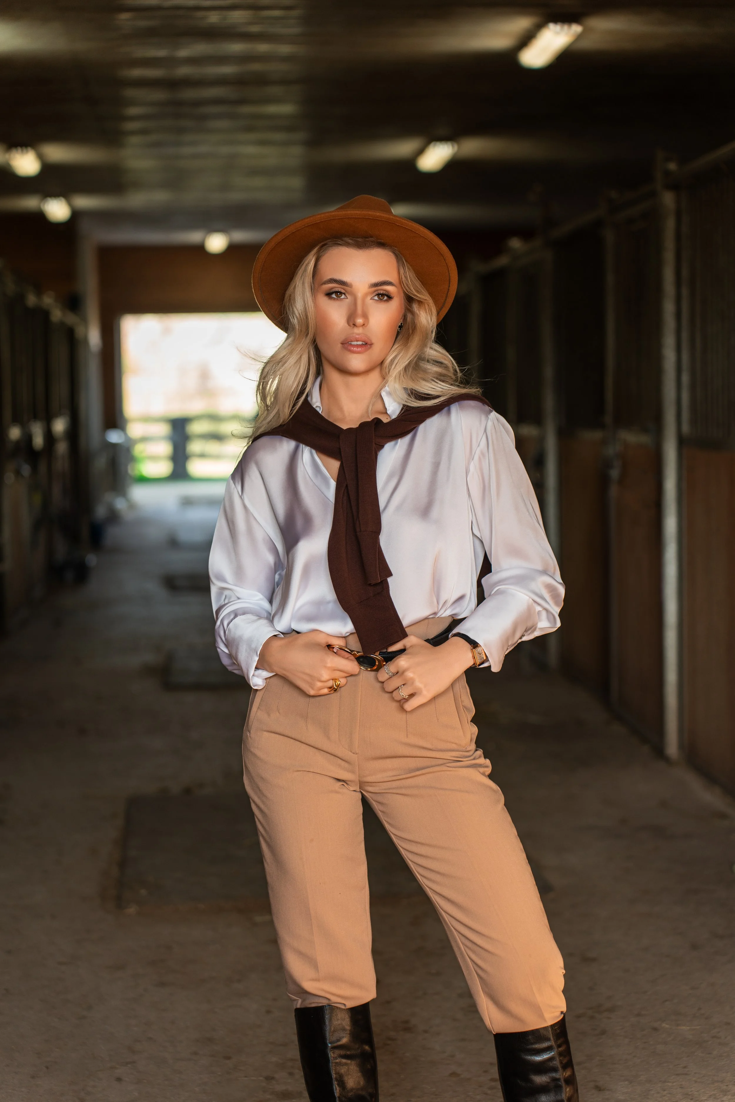 Fashionable woman in a stable wearing a hat, light blouse, beige pants, scarf, and boots.