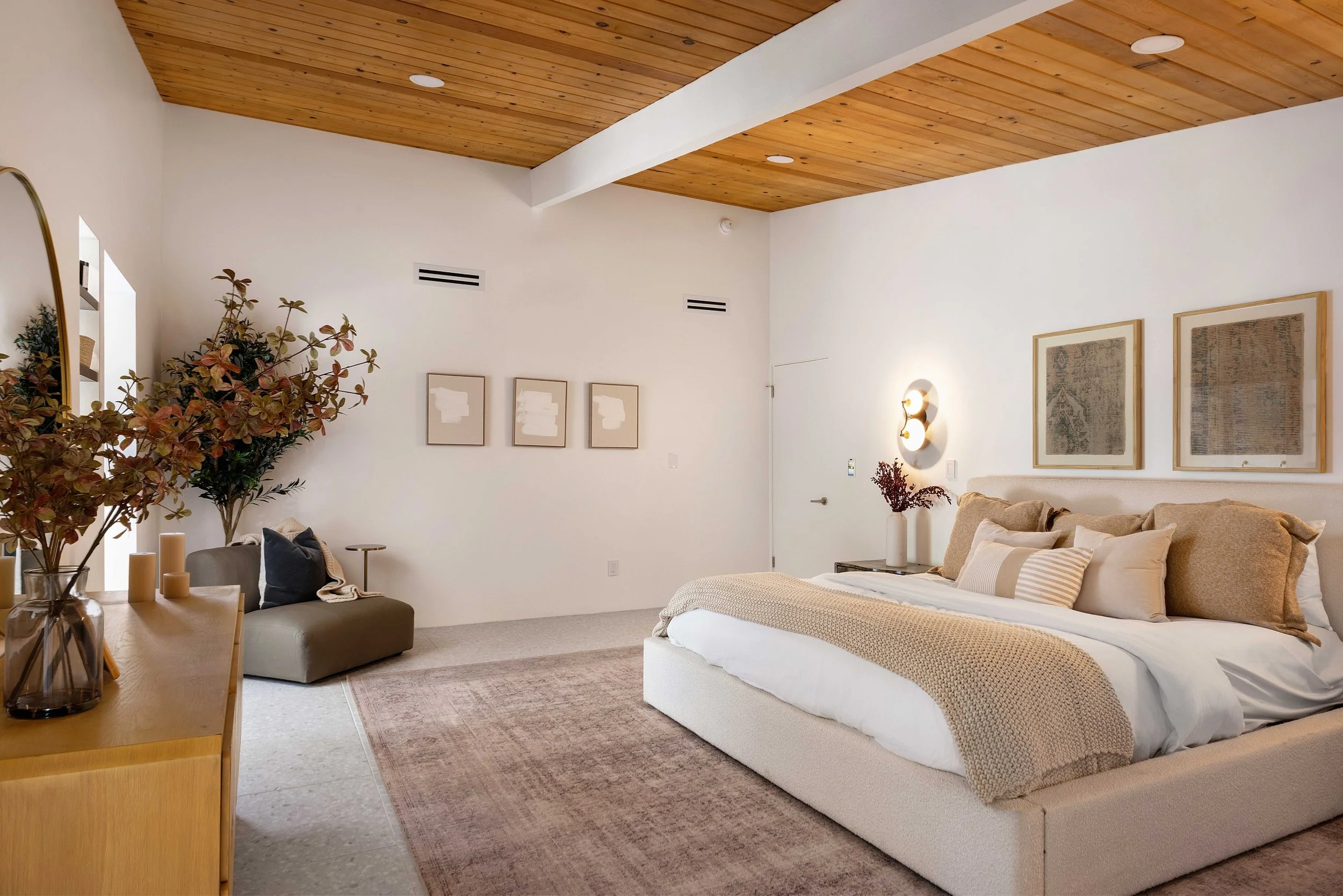 Modern bedroom with a wooden ceiling, large bed, beige bedding, decorative pillows, wall art, vase with plants, and ambient lighting.