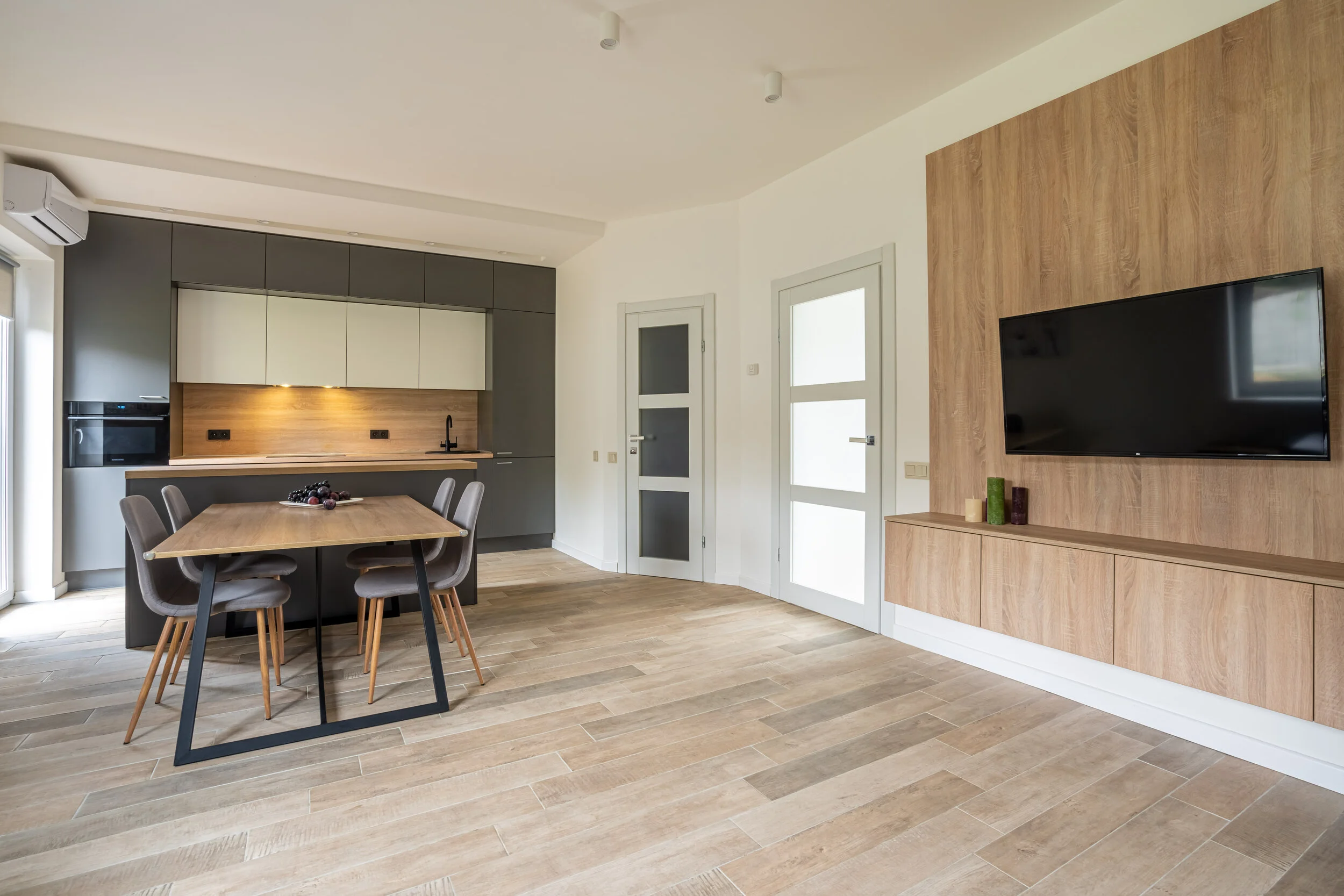 Modern kitchen and living area with light wood flooring, gray and white cabinetry, a wooden dining table with four gray chairs, a flat-screen TV on a wooden accent wall, and a decorative wall cabinet.