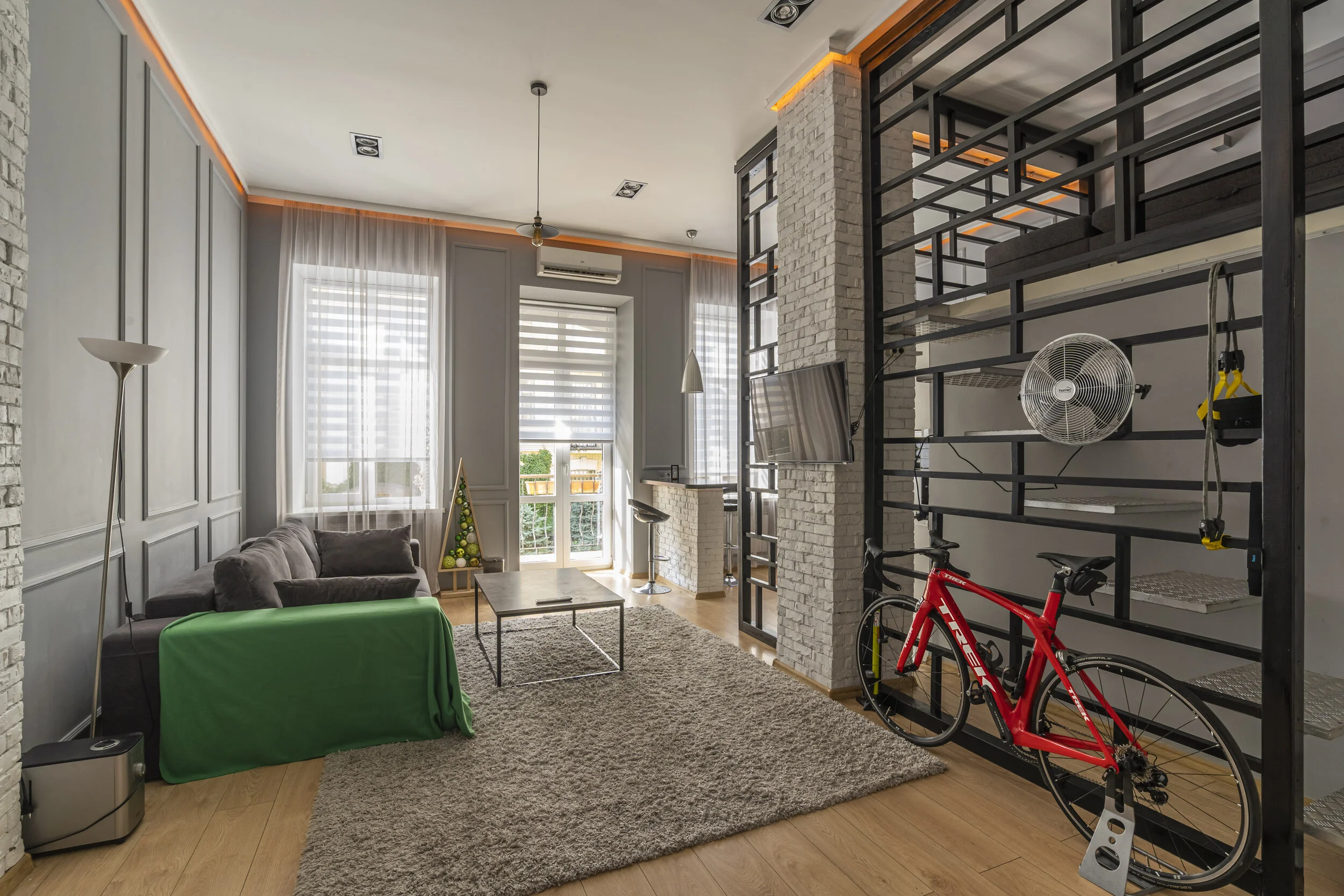Modern loft-style living room with a gray sofa, green throw, coffee table, and plush rug. Features a red bicycle on a stand, wall-mounted TV, loft bed, and brick accent walls. Large windows with blinds allow natural light.
