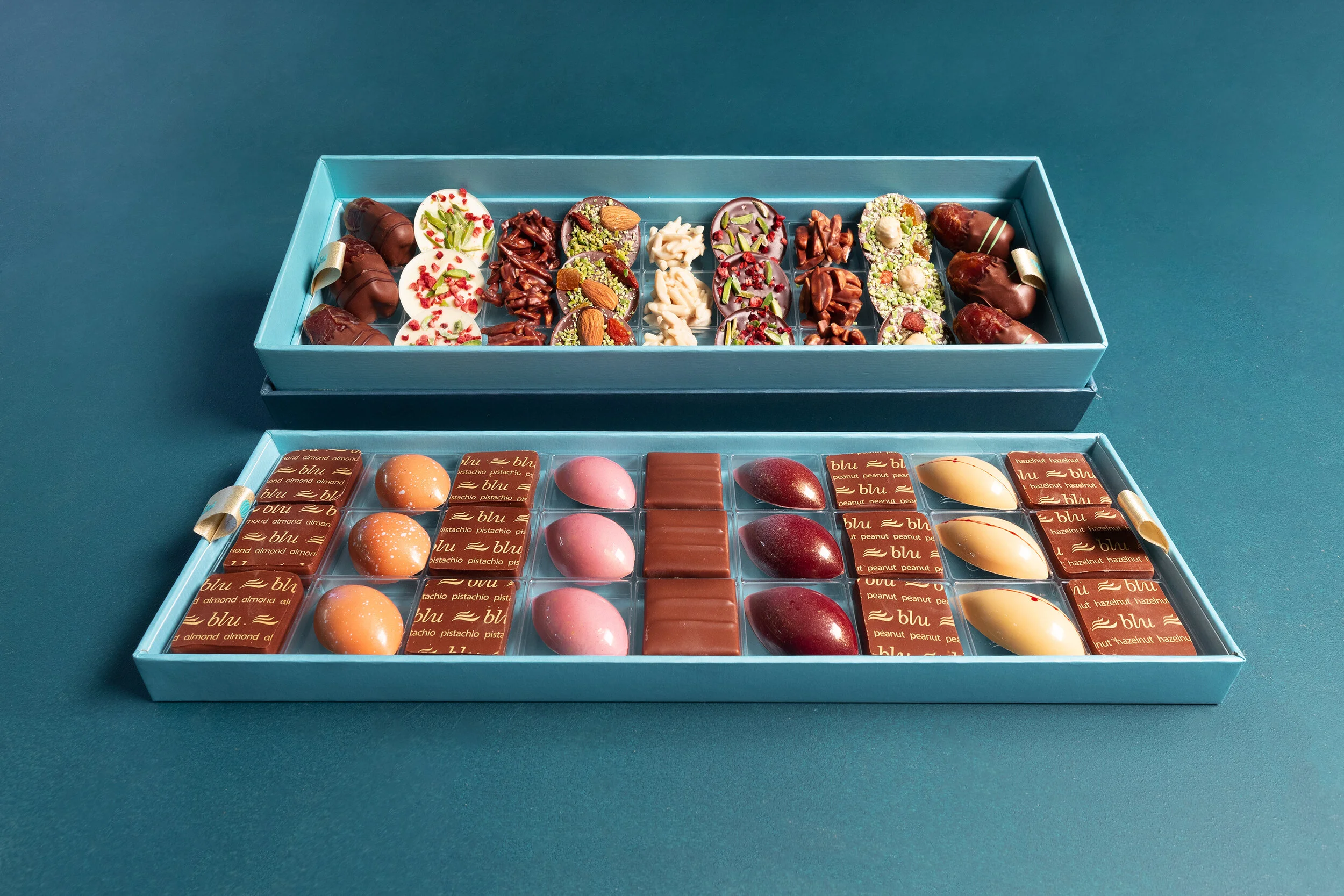 An open box displaying an assortment of handcrafted chocolates with various toppings and fillings, arranged neatly in two layers on a teal background.