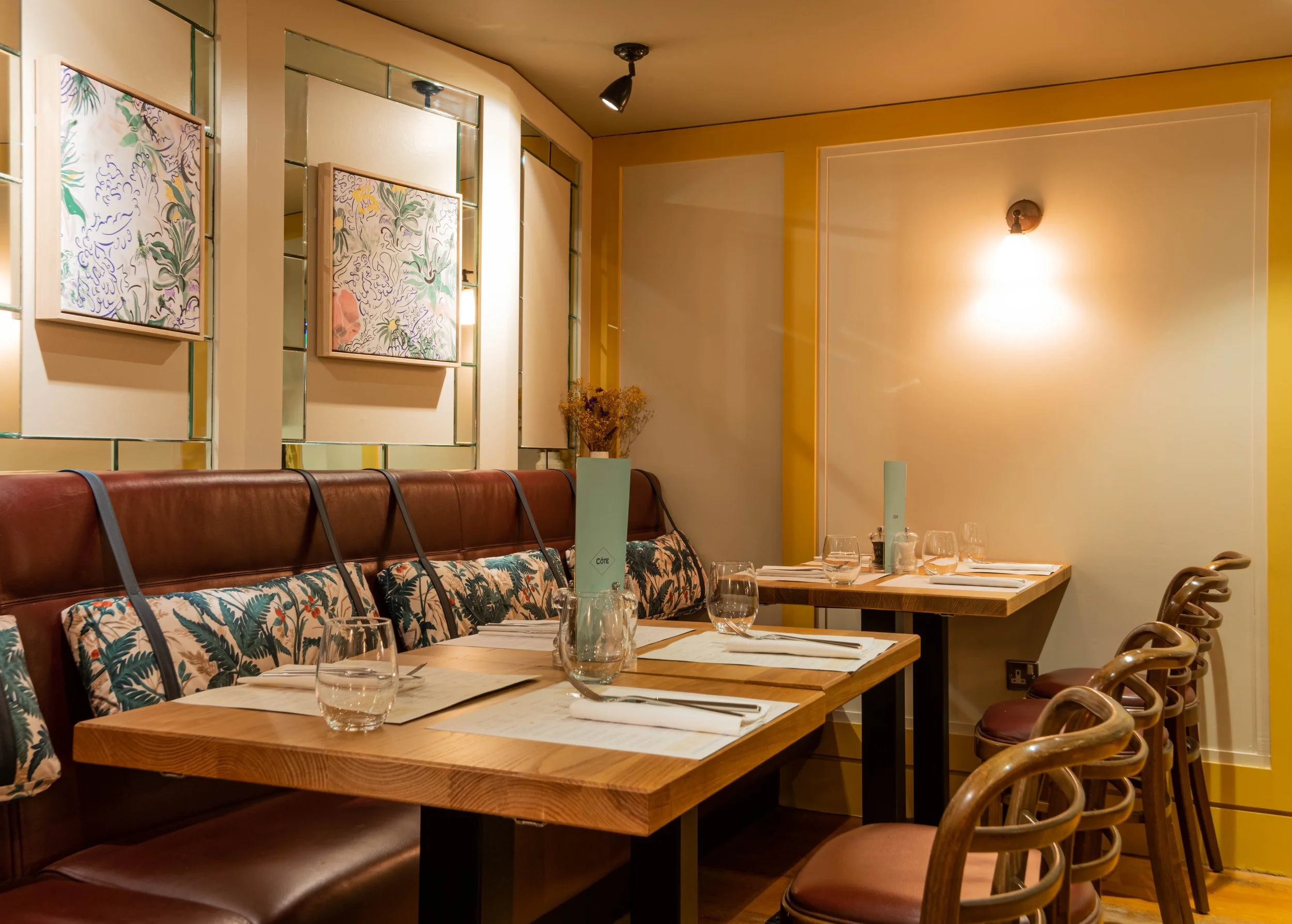 Cozy restaurant interior with wooden tables, leather bench seating, decorative pillows, and chairs. Tables are set with glassware, utensils, and menus. The walls feature abstract floral artwork and warm lighting.