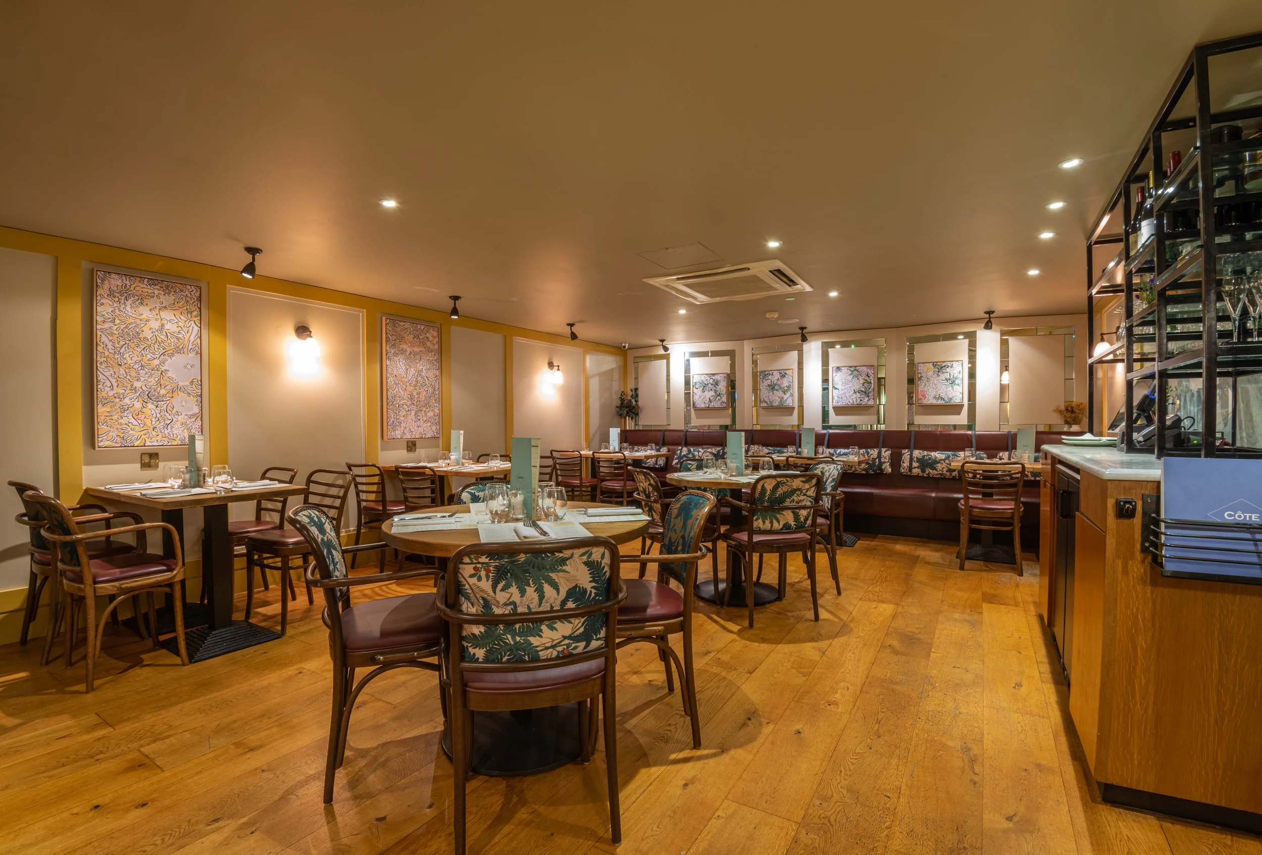 Interior of a modern restaurant with wooden flooring, round tables, and chairs. The walls feature decorative artwork and subtle lighting. The space is elegant and neatly arranged for dining.
